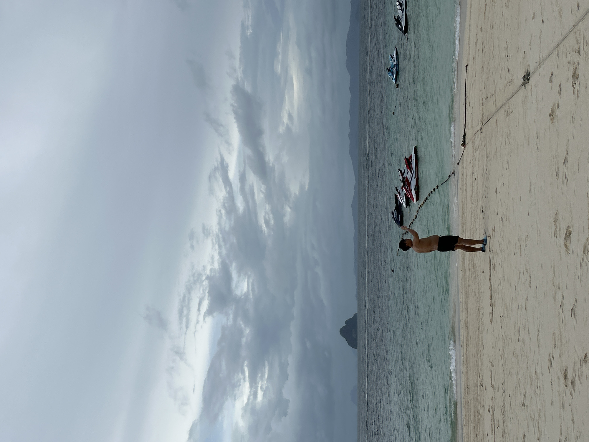 A man stands on a sandy beach in Tambon Phru Nai, Thailand, holding a rope that extends into the water. The sky is overcast with gray clouds. The ocean is a muted blue-green, with gentle waves lapping at the shore. Several jet skis are visible in the water, some clustered together and others more spread out. In the distance, a rocky island with a pointed peak rises from the sea, and a hazy mountain range forms the horizon. The beach is made of light-colored sand with some footprints visible. A thick rope is anchored to the sand and stretches out into the water, possibly to moor the jet skis.