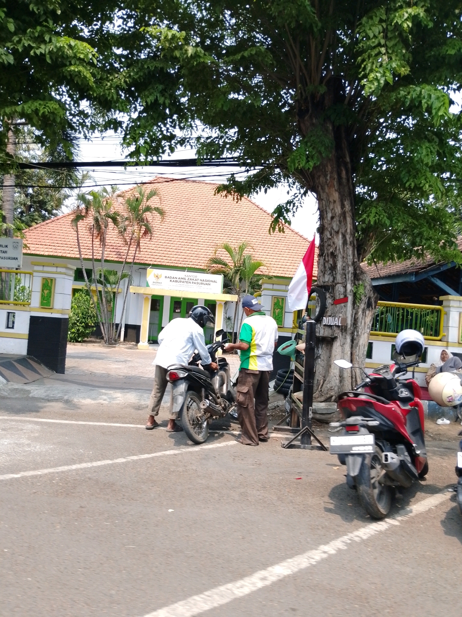 In front of the Badan Amil Zakat Nasional (BAZNAS) building in Pasuruan, Indonesia, two men are interacting with motorcycles. One man, wearing a white shirt and brown pants, is standing next to a black motorcycle, appearing to be working on it. He has a helmet on his head. Another man, in a green and white shirt and brown pants, is also near the motorcycle, and he is wearing a blue cap. To the right, a red scooter is parked, with a white helmet resting on its seat. Further to the right, a woman in a headscarf is seated, seemingly observing the scene. A Indonesian flag is visible on a tree trunk behind the men. The BAZNAS building has an orange tiled roof and white walls with green accents. The environment is outdoors, with trees and a paved road.