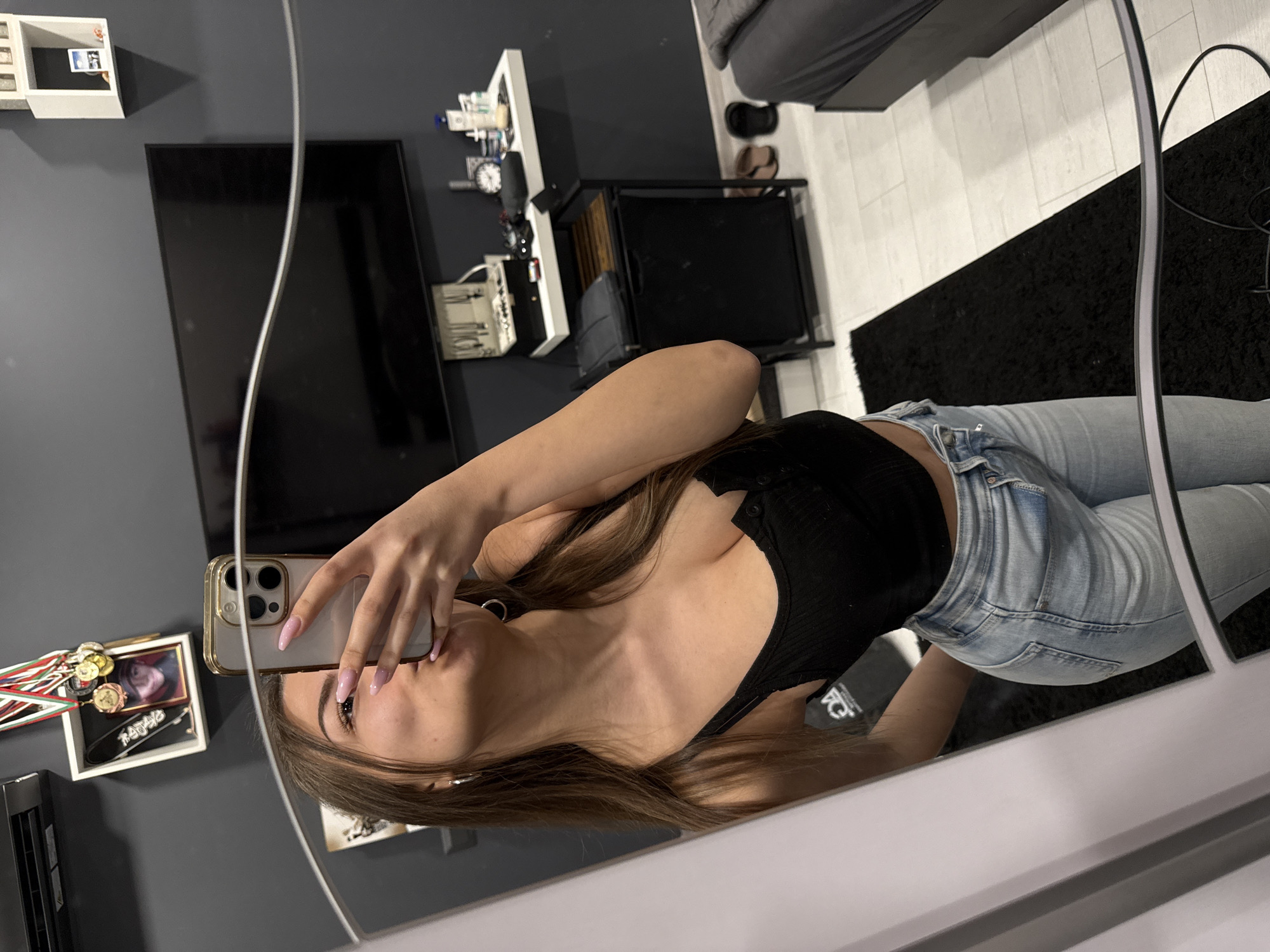 A young woman is taking a selfie in a mirror. The location appears to be a bedroom. She is wearing a black tank top and light blue jeans. The room contains a television, a shelf with various items, a small table, a bed, and a dark rug. There is no information in the image to indicate that the location is Sofia, Bulgaria.