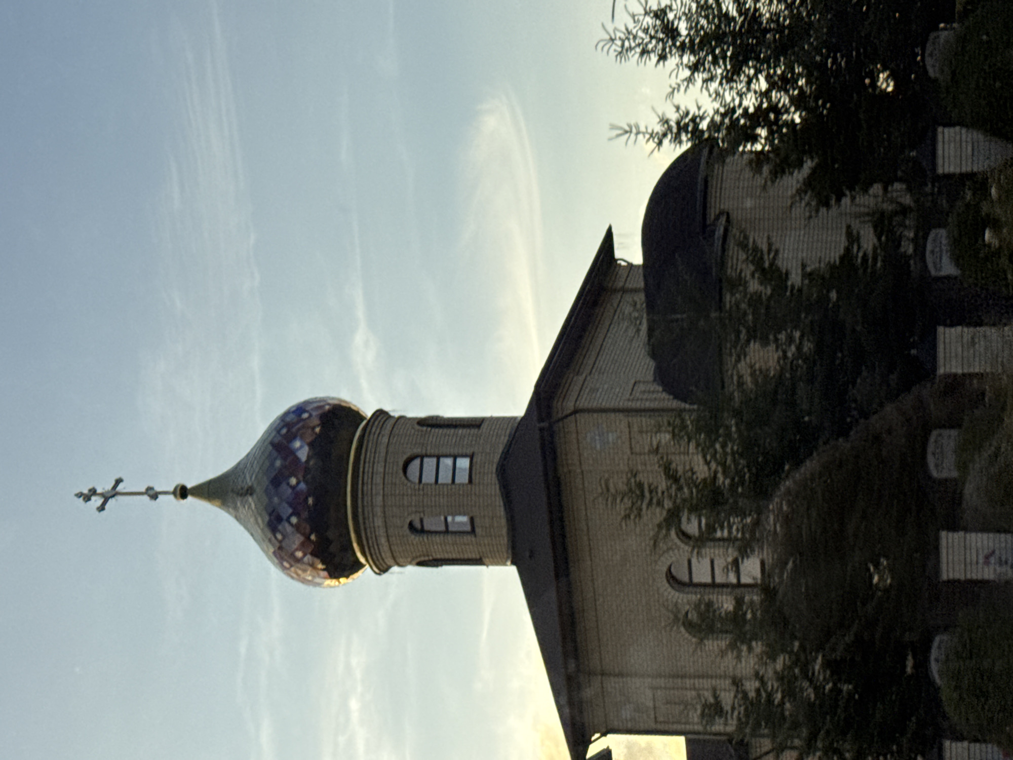 An exterior, medium shot captures an Orthodox church in Mikhaylovsk, Russia. The church is constructed of light-colored brick and features a multi-faceted dome with a cross at its apex. The dome is adorned with various shades of blue and purple, with gold accents. Arched windows are visible on the cylindrical tower beneath the dome, and the main body of the church also has arched windows. Dark green coniferous trees frame the church on both the left and right sides. The sky above is a pale blue with wispy, white clouds. The lighting suggests it is late afternoon or early evening, with a warm glow on the dome. The foreground is partially obscured by dark foliage and the tops of white brick pillars.