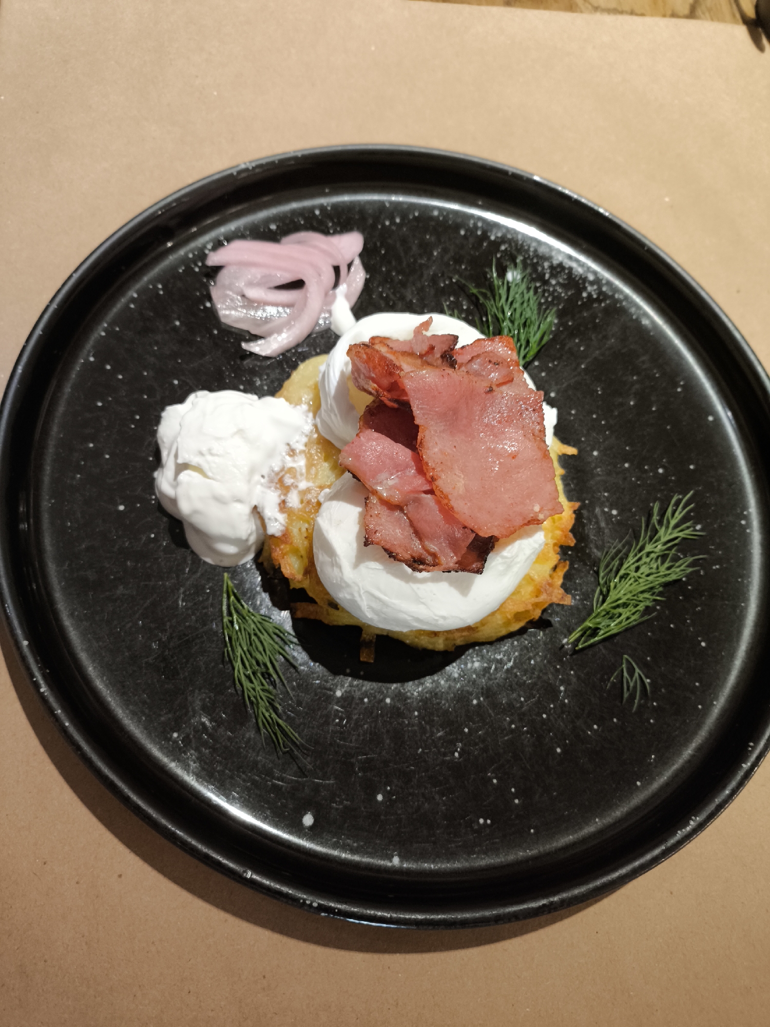 A top-down, slightly angled image depicts a savory dish centered on a dark, speckled ceramic plate. The main subject consists of at least two golden-brown, pan-fried potato pancakes (draniki) forming a base. These are topped with two distinct portions of a creamy white substance, likely sour cream or crème fraîche; one large dollop is positioned to the left of the pancakes, and another smaller dollop is directly on top of them. Arranged over the latter white topping are multiple slices of cooked, reddish-pink meat, possibly cured bacon or pastrami, showing some crisping on the edges. To the upper left of the dish, a small cluster of thinly sliced, translucent pink pickled red onions is present. Several sprigs of fresh green dill are used as garnish, with larger pieces placed on the lower left and right of the plate. The black plate features a slightly raised rim and exhibits numerous small white speckles, along with a few larger white spots, which could be part of the glaze or scattered ingredients. The plate rests on a plain, light brown, matte surface, consistent with a wooden table or a large sheet of craft paper, under soft, diffuse indoor lighting. This scene is situated in Izhevsk, Russia.