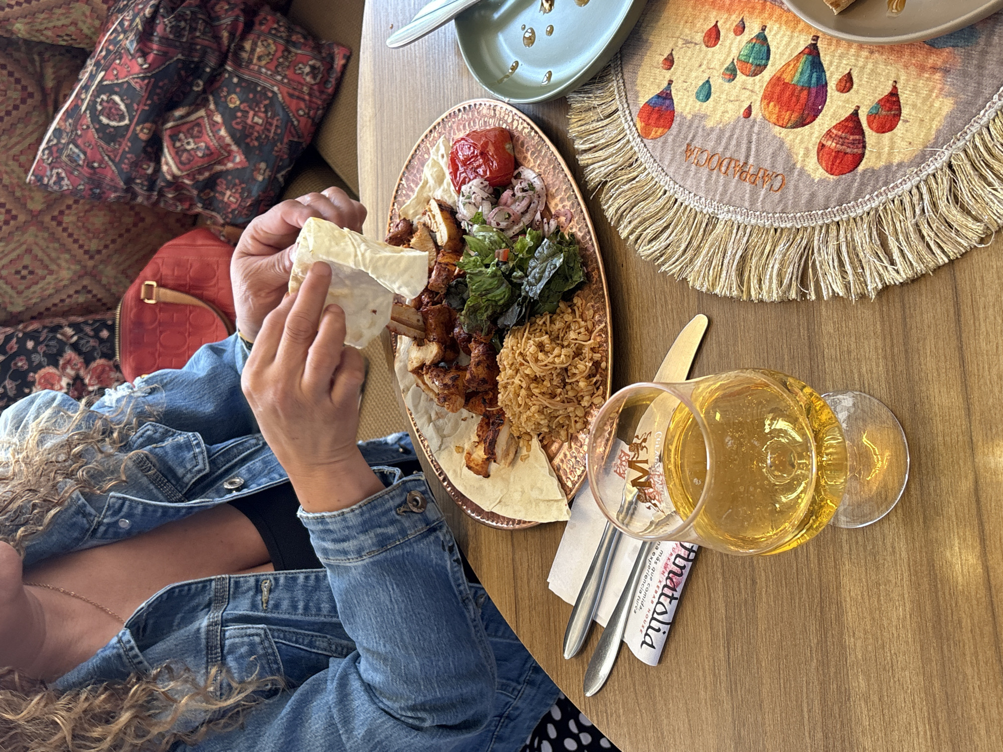 An indoor dining scene is depicted at "Anatolia Turan Kebab House" in Heroica Puebla de Zaragoza, Mexico. A person, partially visible with curly blonde hair and wearing a denim jacket, is actively preparing a meal. Their hands are shown folding a piece of flatbread around food from a large, hammered copper-colored plate. The plate holds grilled meat, rice pilaf, a small green salad with red onions, a roasted tomato, and additional flatbread. A stemmed glass containing an amber liquid rests on the wooden table. A set of cutlery is placed on a white napkin bearing the restaurant's name. To the right, an empty green plate shows food residue. A semicircular placemat with fringes displays colorful hot air balloons and the word "CAPPADOCIA." In the background, a red Carolina Herrera handbag is visible on upholstered seating adorned with patterned cushions.