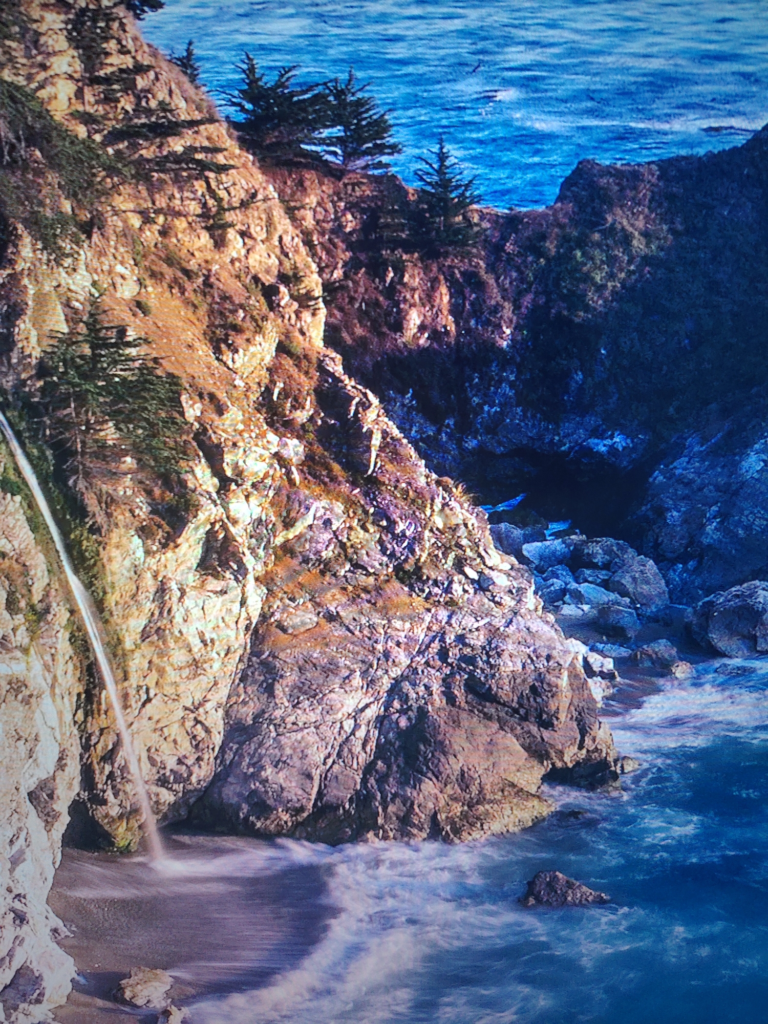 This image depicts McWay Falls in Julia Pfeiffer Burns State Park, California. The scene features a rocky coastline with a waterfall cascading directly onto the beach. The cliffs are covered in vegetation including evergreen trees. The ocean is visible in the background and foreground, with waves crashing onto the shore and rocks.