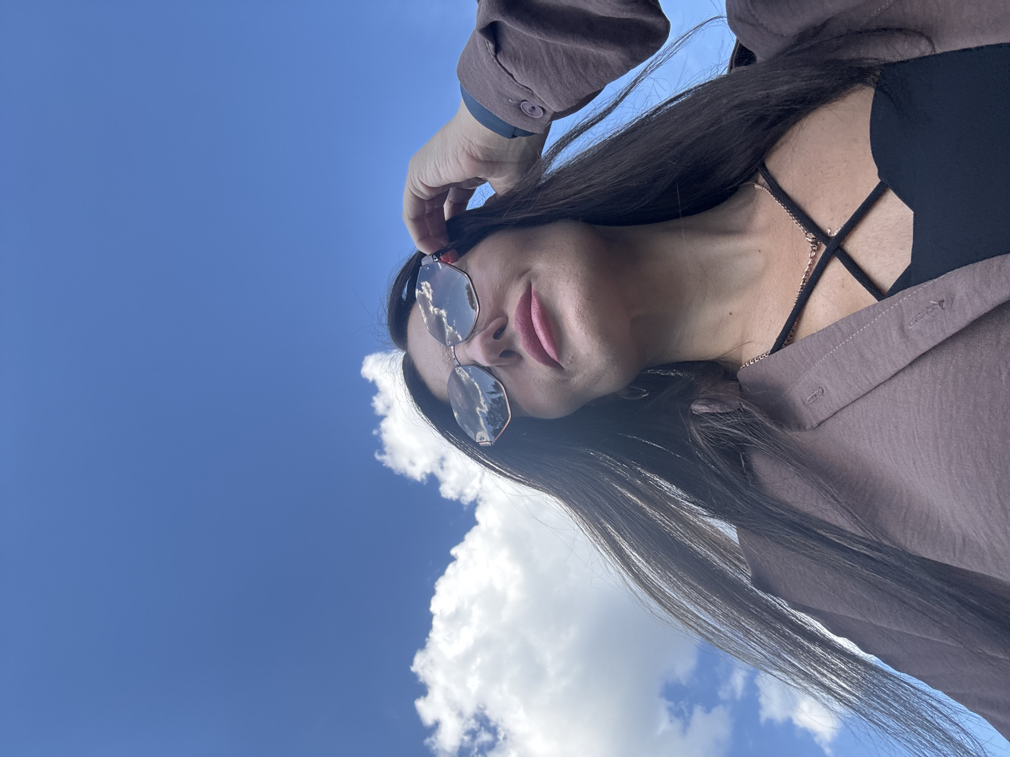 A low-angle, outdoor, waist-up shot shows a woman wearing sunglasses and a light brown jacket over a black top. Her dark hair is long and flowing. She is looking up and adjusting her sunglasses with her right hand. The sky is blue with some white clouds. The image does not provide any information about the location in the city of Chaikovsky, Russia.