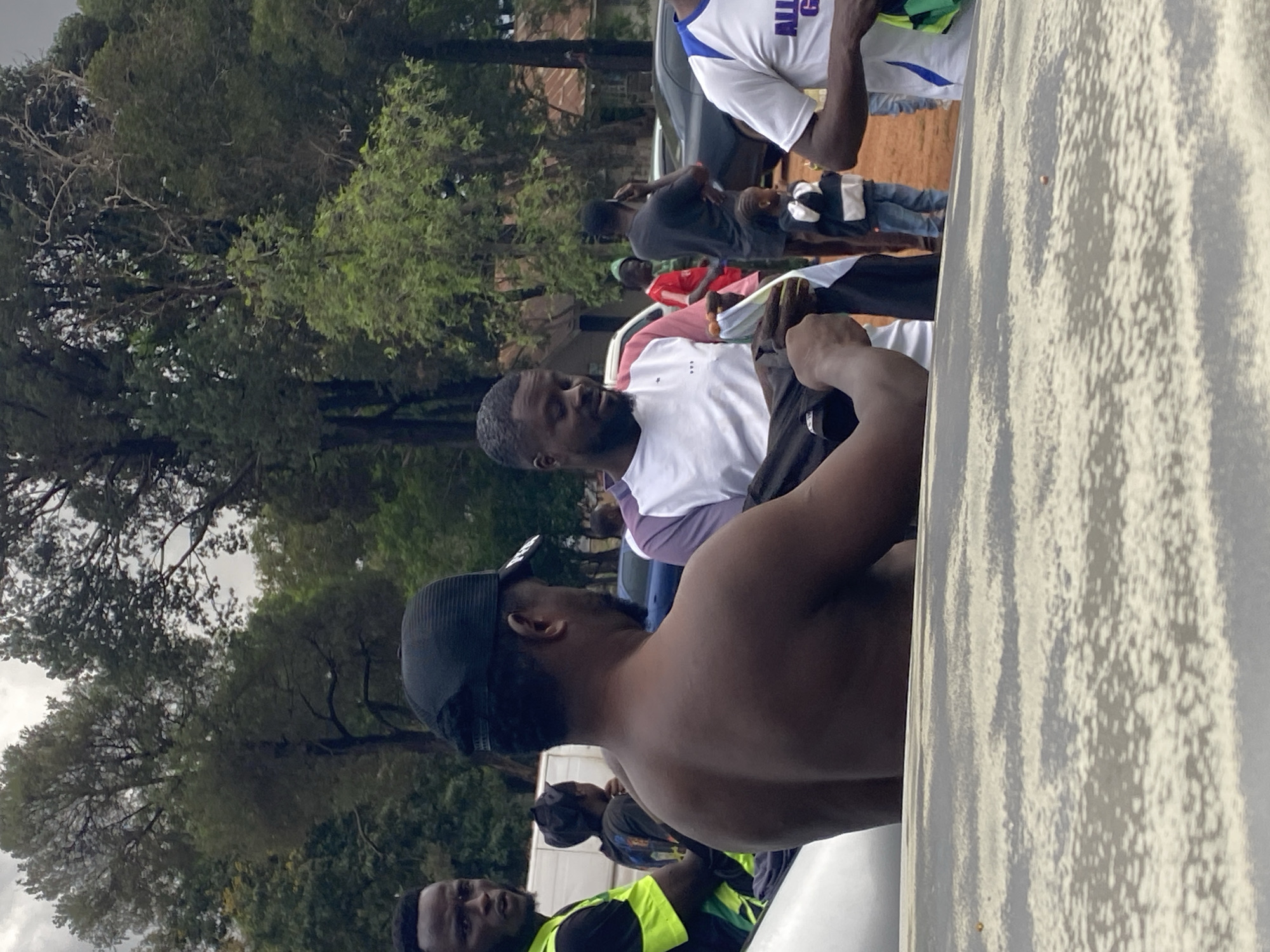 This image, captured outdoors, depicts a gathering of several people under what appears to be an overcast sky. The photo is taken from a low angle, looking across a white, textured surface that dominates the right foreground, possibly the side of a large vehicle or structure.
In the immediate foreground, a shirtless man wearing a black baseball cap is seen from behind and his right side, facing towards the left of the frame. Just to his left and slightly behind, another man with a beard, dressed in a white short-sleeved shirt, looks towards the left, holding or observing papers in his hands with a neutral expression.
Further back, towards the left, several other individuals are visible. A person wearing a white shirt with purple accents is partially seen, also appearing to handle documents. Near them, a person in a dark t-shirt and jeans stands with a child beside them, who is wearing a white and black outfit. In the bottom left corner, a man in a neon yellow/green safety vest is visible, looking towards the right, alongside another man in a dark t-shirt and cap. A dark-colored car is parked in the mid-ground, behind some of the individuals.
The background is filled with dense green trees and foliage, suggesting a natural or semi-urban setting. The ground visible through the crowd appears to be reddish-brown earth. The diffused lighting indicates an overcast day or that the scene is in shade. On the white and purple shirt, the partial text "ALL G" is discernible. The overall scene suggests a casual assembly or an informal event, possibly involving the distribution or review of documents, set in an outdoor area in Jos, Nigeria.