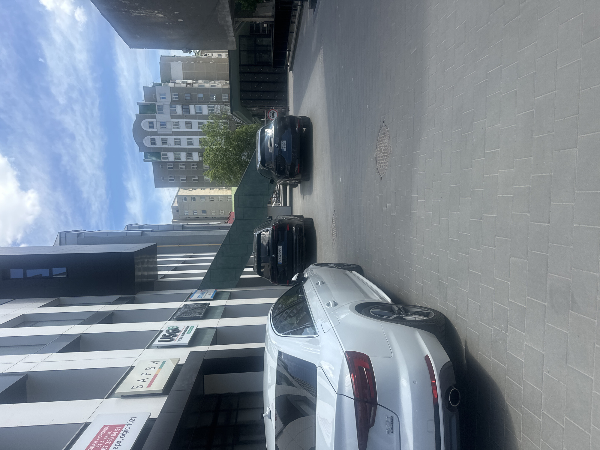 The image shows a street scene in what appears to be an urban area in Ternopil, Ukraine. In the foreground, a white Audi car is parked next to a modern building. The building has a sign that reads "Барви", which translates to "Colors" in Ukrainian. There are other signs visible on the building including "USGO" and "VIENNA GRANT GROUP". The sign next to the building states that the property is for rent (ОДАЖ ОРЕНДА) and is 57.4 square meters, office 1021. Further down the street, two dark-colored BMW vehicles are parked. License plates are visible on the cars with "BO 3222 BC" being the number on one car and "8104" visible on the other. The street itself is paved with grey rectangular bricks. In the background, there are apartment buildings of varying heights and designs under a partly cloudy sky.