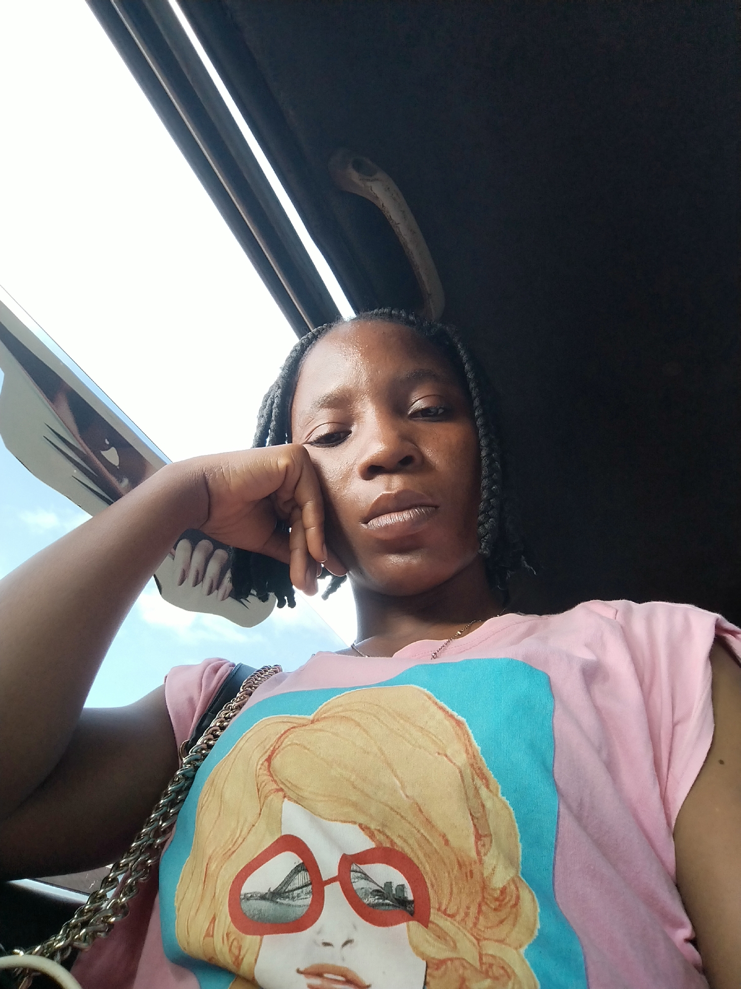 This is a close-up, waist-up, outdoor selfie of a young woman with dark skin and braided hair, wearing a pink t-shirt with a graphic print of a woman wearing red-rimmed sunglasses. She is resting her chin on her hand, looking towards the camera with a neutral expression. The background suggests she is inside a vehicle, possibly a bus or car, with the sky and some structures visible through a window. The overall lighting is bright and natural, indicating it is daytime. The setting is likely Benin City, Nigeria, given the user's prompt, though no specific landmarks are identifiable to confirm this.