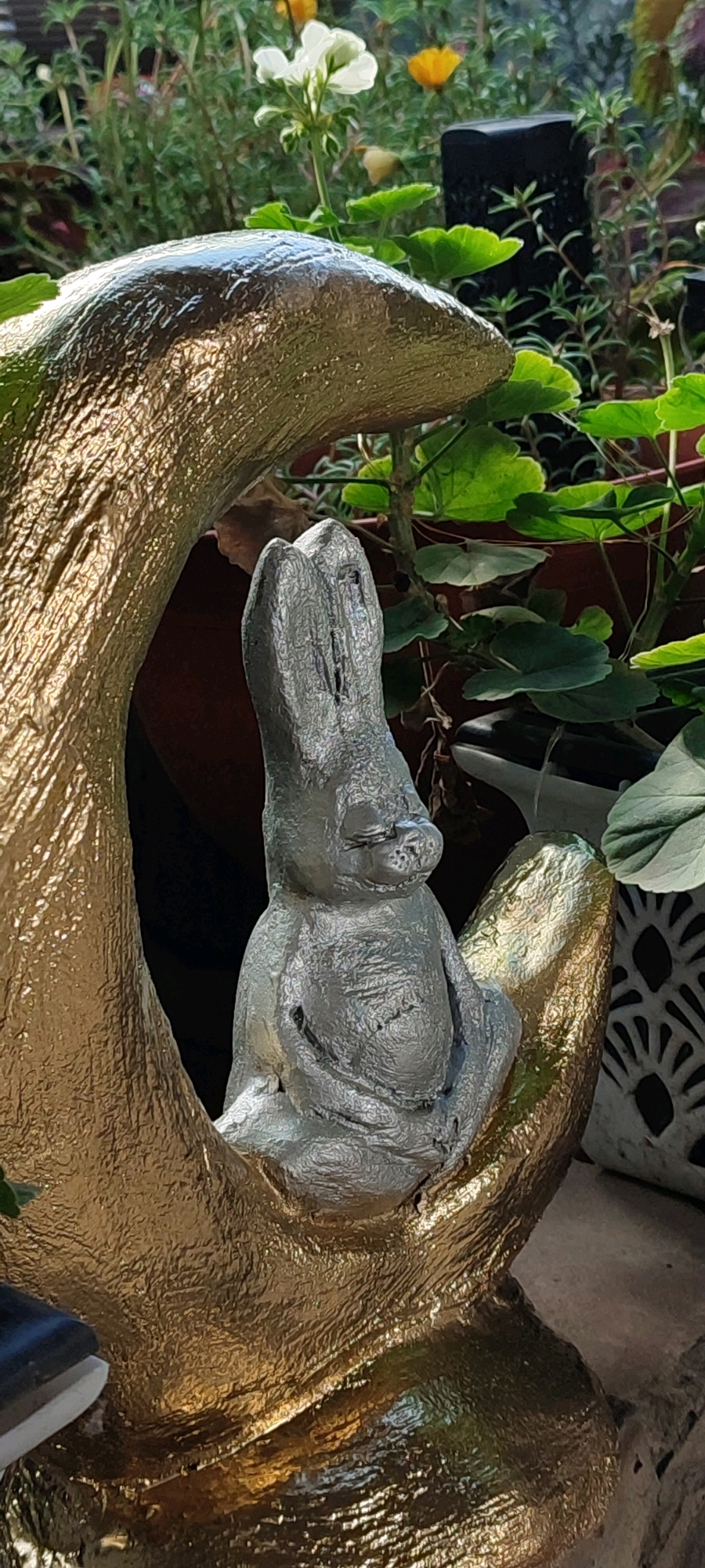 A statue of a grey, cartoonish rabbit with closed eyes and a small nose is centered in the frame. The rabbit is sitting within a golden crescent-shaped structure that appears to be made of a textured material, possibly concrete painted gold. The rabbit's ears are long and upright, and its body is rounded. It is leaning back slightly, with its arms resting on its lap. The background consists of lush green foliage and plants in pots, suggesting an outdoor setting. In the distance, there are glimpses of yellow and white flowers. To the right of the statue, a decorative white planter with an intricate pattern is visible. The overall scene appears to be a garden decoration. The image was captured in the city of Belaya Kalitva, Russia.