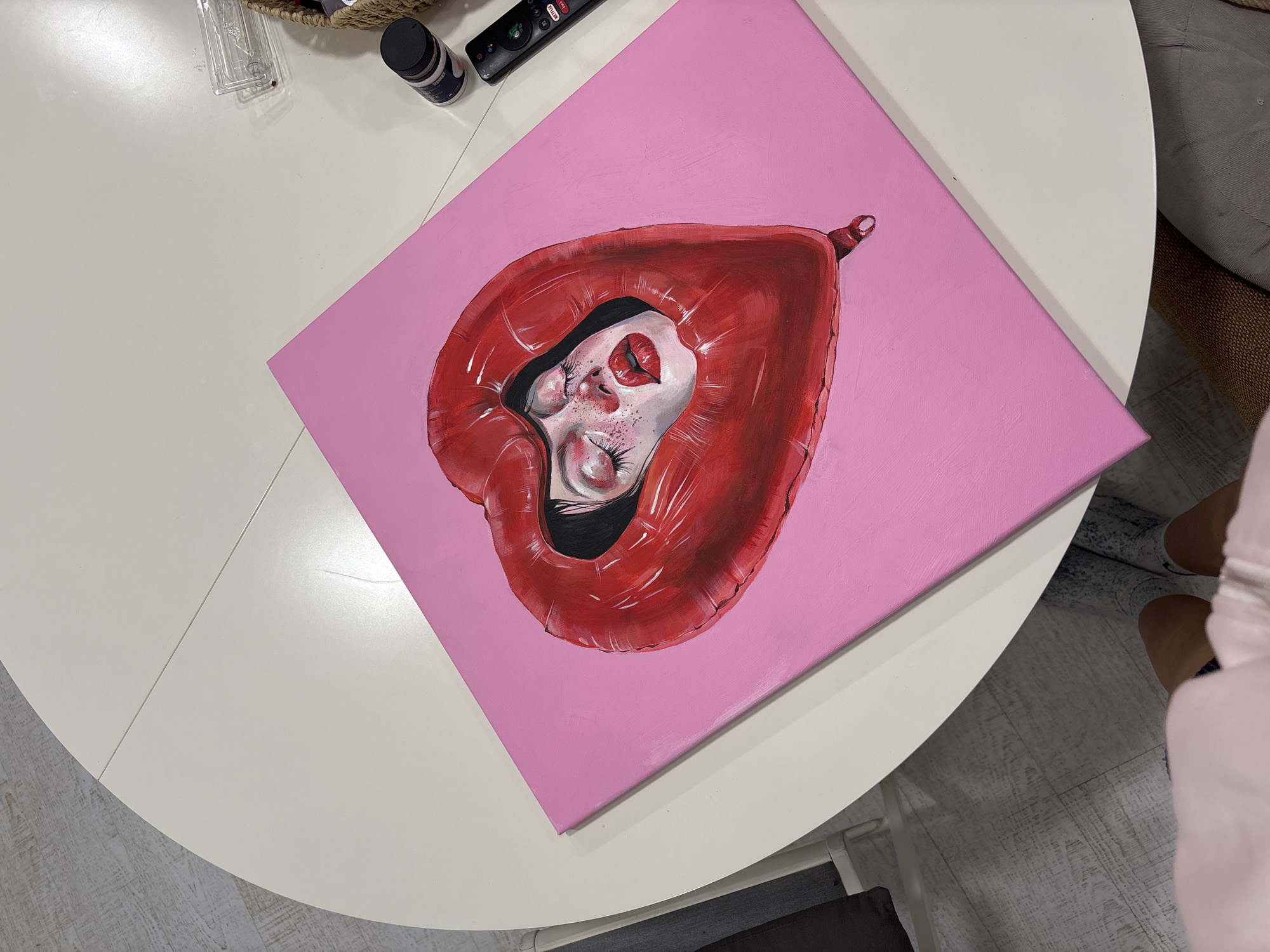 The image shows an oil painting on a pink canvas. The painting depicts a face encased within a red, heart-shaped balloon. The subject's skin tone is a pale white with freckles, and their lips are painted as a small red heart. The painting rests on a white, round tabletop. Adjacent to the canvas are a black jar, a black remote control, and a small wicker basket containing a clear plastic case. A portion of a gray upholstered chair or sofa is visible in the background, along with a person's legs wearing tan shoes. The environment is a room with light-colored flooring. The location is not discernible from the image.