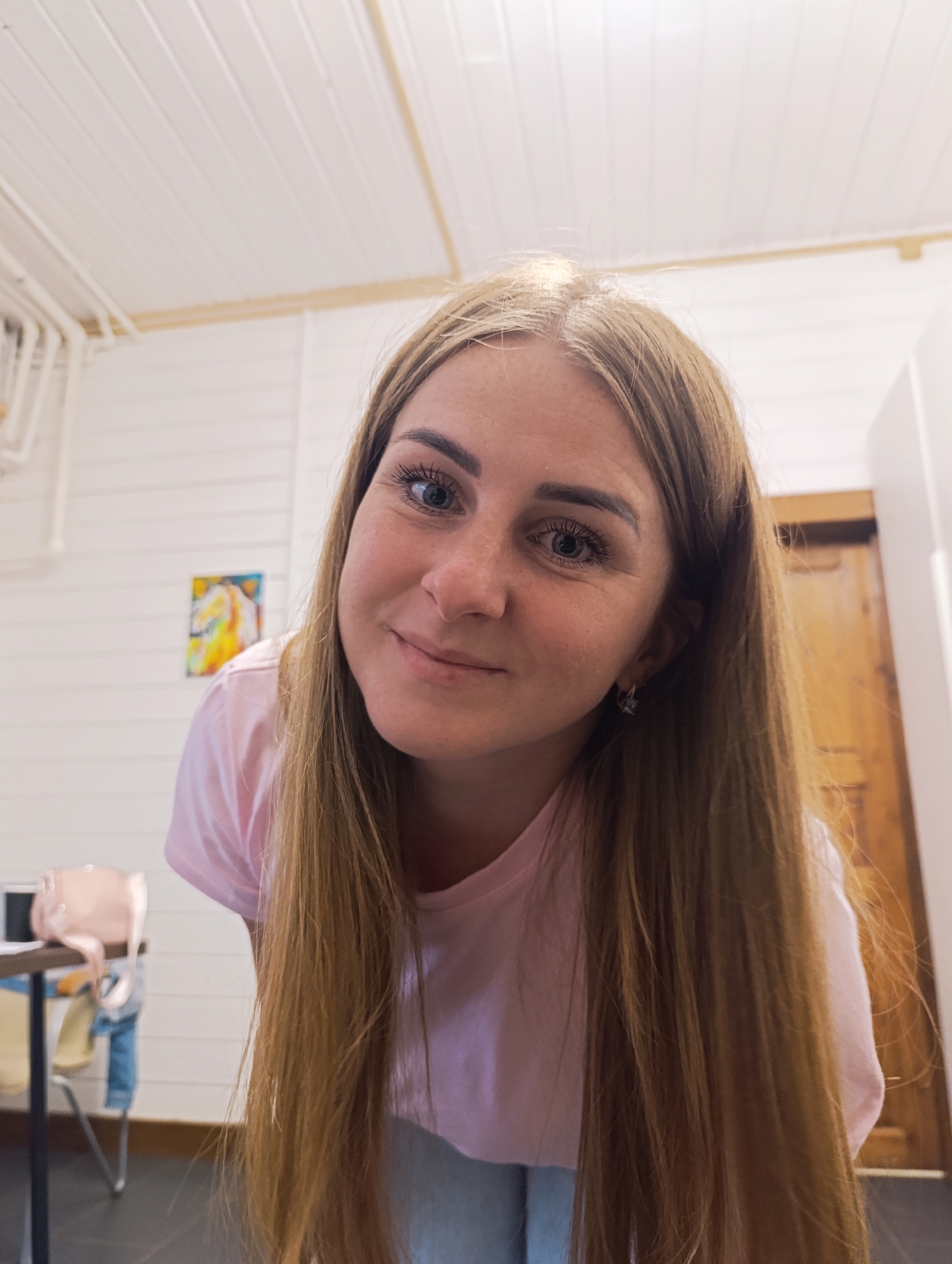 A medium shot captures a young, fair-skinned woman from the shoulders up, looking directly at the camera. She has long, straight, light brown hair that falls around her shoulders and chest. Her eyes are a light blue-gray, and she is wearing mascara. She has a slight smile, and her face is lit from above. She is wearing a light pink t-shirt. In the background, a white wall with horizontal planks is visible, along with a light wood door on the right. On the left, a small, framed artwork with yellow and orange hues hangs on the wall. Further left, a table and chairs are partially visible, with a pale pink bag resting on the table. The ceiling above is white with visible wooden beams and what appear to be recessed lights. The overall impression is a casual, indoor setting.