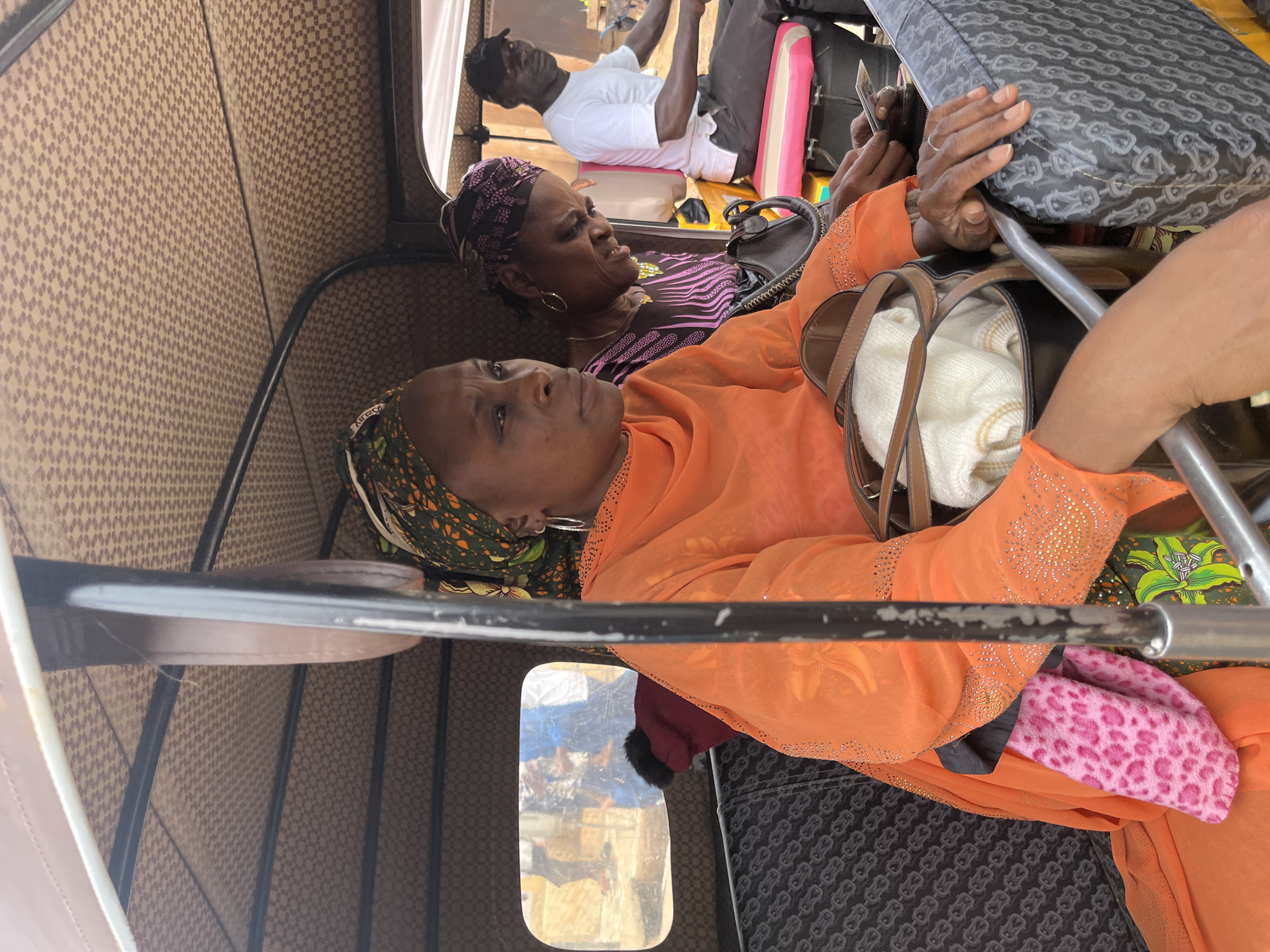 The image shows the interior of a vehicle, likely a public transport auto-rickshaw, in Gura Top, Nigeria. Two women are visible as passengers. The woman in the foreground wears an orange garment and a headscarf with green and brown floral patterns. She is holding onto a metal bar and has a handbag beside her containing a white object. The second woman, sitting behind the first, wears a purple patterned top and a dark headscarf. She is gazing towards the right. In the background, the driver of the auto-rickshaw is partially visible wearing a white t-shirt. Luggage items, including a pink and black case, are present. The interior of the auto-rickshaw has a patterned brown and gray design. Through a window, a glimpse of the city environment is visible.