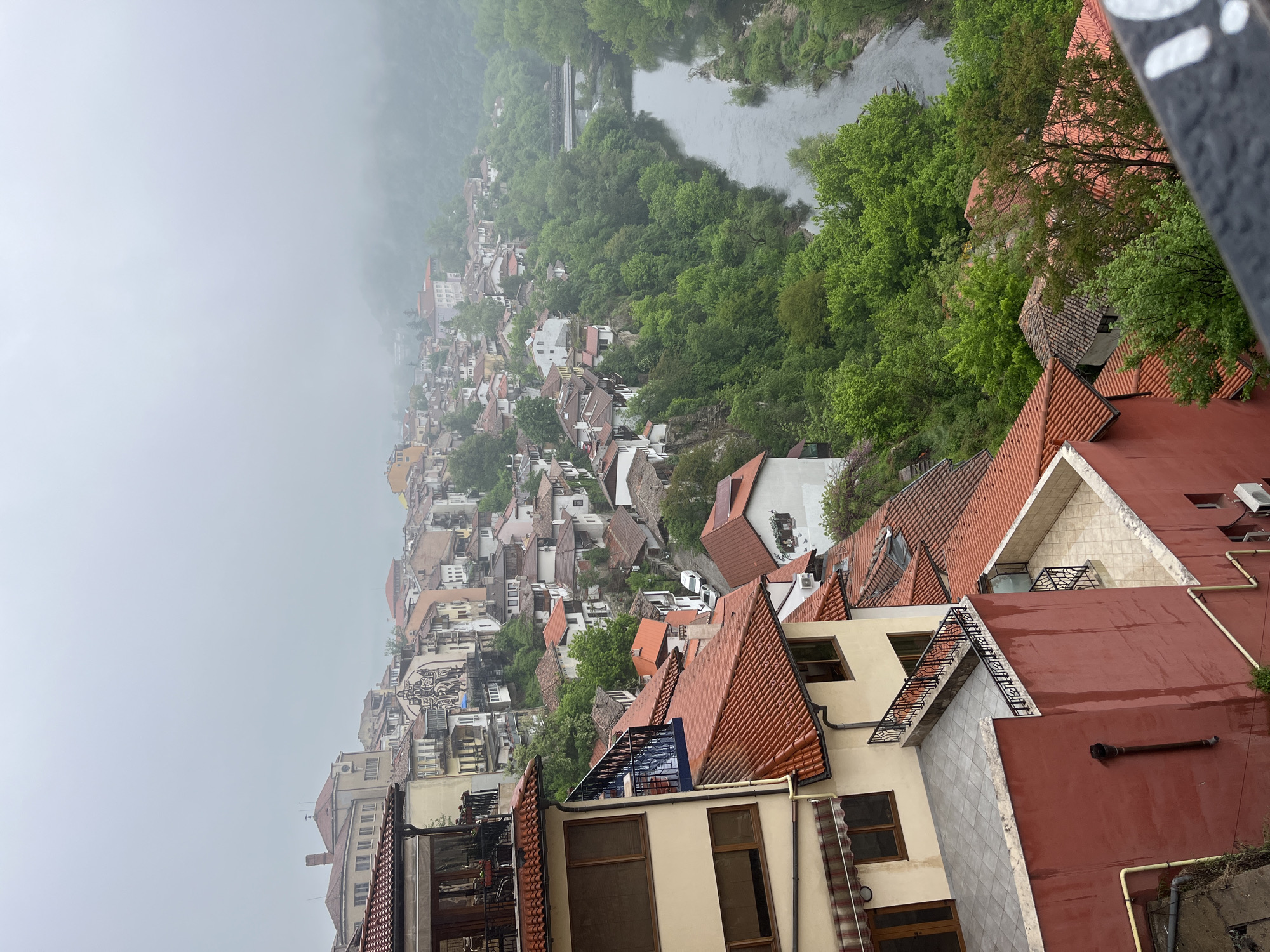 This photograph, taken from a high vantage point, depicts a section of the city of Veliko Tarnovo, Bulgaria, on a cloudy or foggy day. The foreground features rooftops of buildings, primarily with orange or red terracotta tiles, and some with modern, flat roofs. There are windows and architectural details visible on the sides of the buildings closest to the camera. In the middle ground, the cityscape slopes downwards, showcasing a dense collection of buildings with varying roof colors and architectural styles. Trees are interspersed among the buildings, adding patches of green to the scene. Further in the background, the fog or mist obscures some of the buildings and landscape. A river snakes through the landscape, bordered by trees. A bridge can be partially seen spanning the river. The overall scene conveys a sense of depth and the historic, layered architecture characteristic of Veliko Tarnovo.