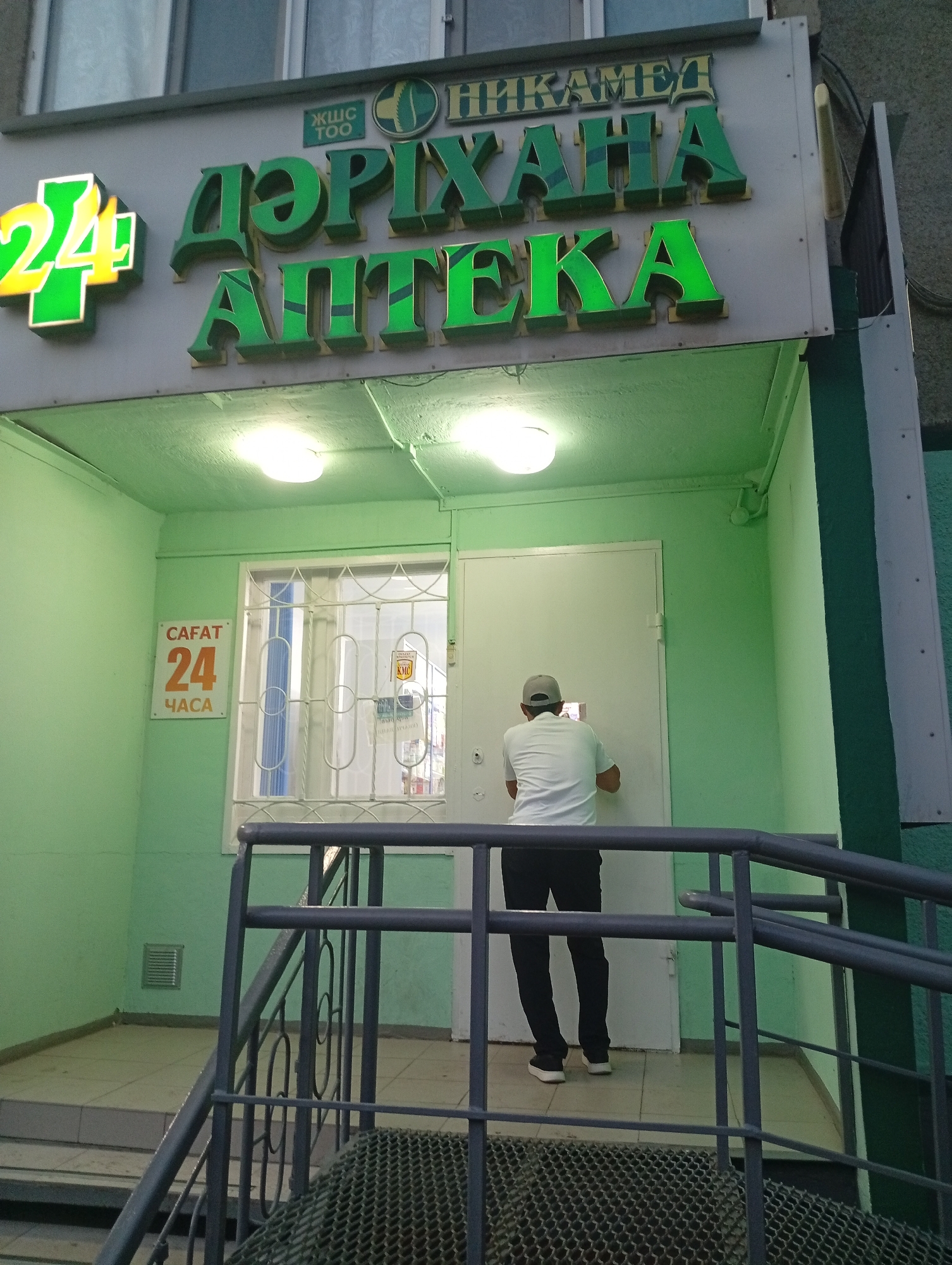 The image shows the exterior of a pharmacy in Oral, Kazakhstan. The pharmacy has a prominent green and yellow sign with the number "24" in a cross shape on the left, and green lettering that reads "DƏRİXANA APTEKA" in the center. Above this, there is smaller text that appears to be a company name or slogan. The pharmacy entrance is painted a light green.
A person wearing a white polo shirt and black pants, with a grey baseball cap, stands with their back to the camera, facing a white door. They appear to be interacting with something on or near the door, possibly a tablet. To the left of the entrance is a window with metal bars, and a sign that reads "САҒАТ 24 ЧАСА" in orange and white text, indicating the pharmacy is open 24 hours. The lighting suggests it could be daytime, but the indoor lighting is also visible, creating a slightly artificial glow. The scene is set on what appears to be a street or sidewalk, with a metal railing and stairs in the foreground, suggesting the entrance is elevated. The overall impression is a commercial establishment operating in a residential or mixed-use area.