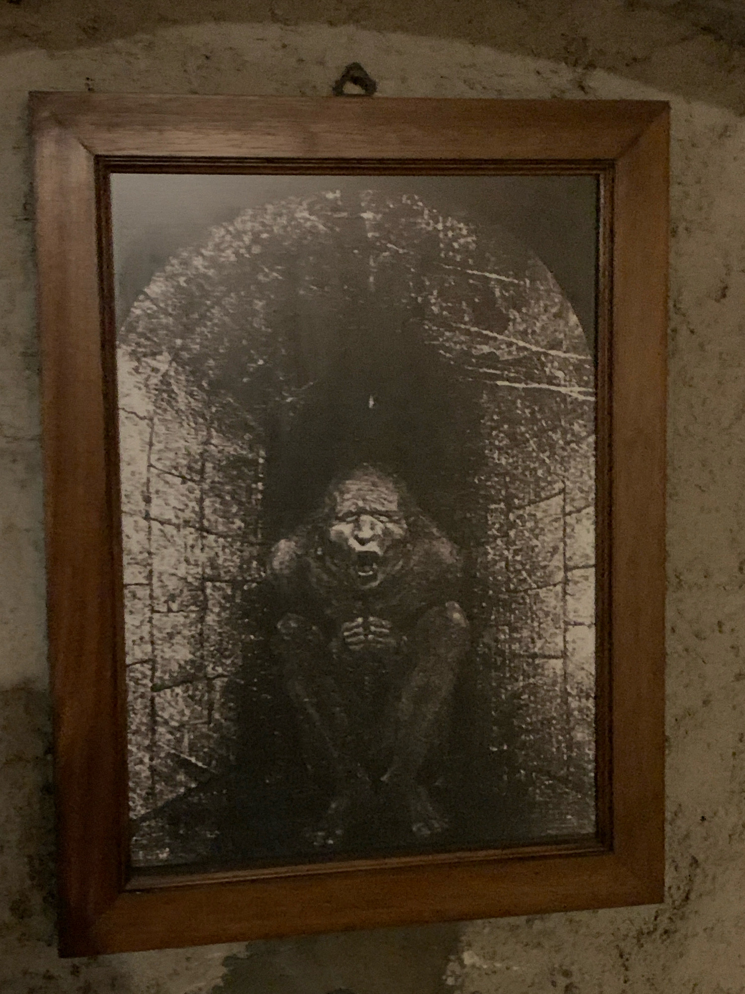 An unframed, black and white photograph depicts a grotesque, hunched-over figure within a dimly lit, brick-lined tunnel or alcove. The figure, possibly a creature or a heavily distorted person, has a wide, open mouth with visible teeth and a contorted facial expression. Its body is crouched, with knees drawn up and hands clasped in front of its chest. The tunnel walls are textured and appear ancient, with a rough, uneven surface. The overall lighting is dark, with shadows obscuring much of the background. The photograph is hung on a textured, stone-like wall in what appears to be an indoor setting. The location is not specified in the city of City of San Marino, San Marino.