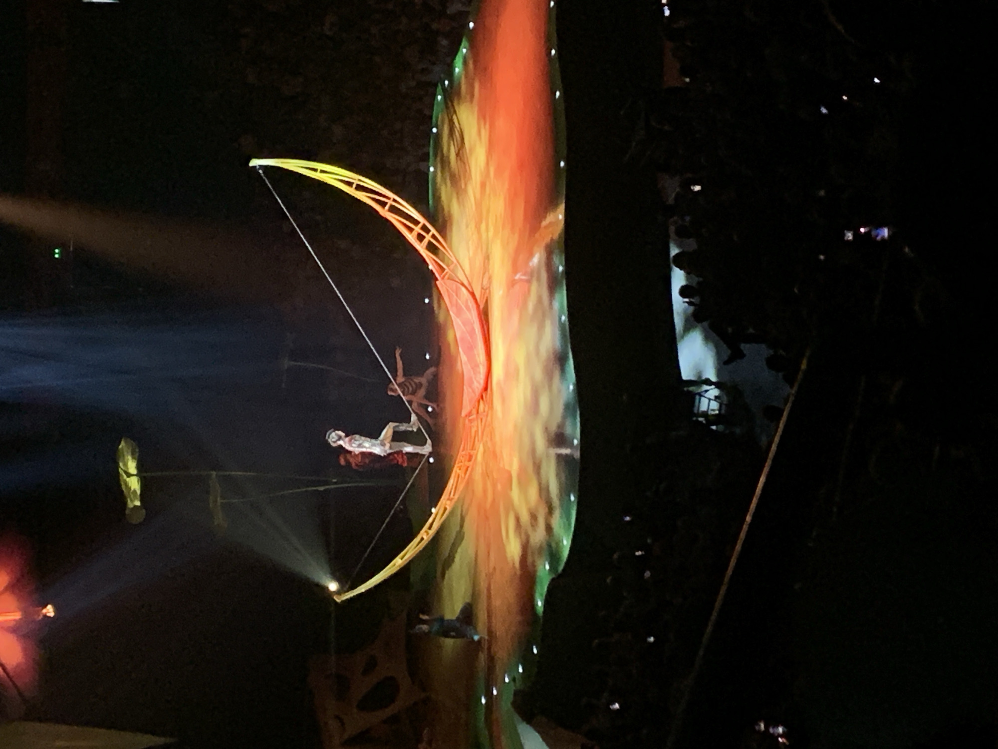 The image captures a Cirque du Soleil performance, likely a show within a larger event, in a dark environment. The performers are engaged in aerial acrobatics using equipment, including a curved, illuminated structure resembling a bow. A large screen displays a fiery, abstract visual backdrop. The audience is visible on the right side of the frame, partially obscured by darkness, with numerous phone screens illuminated as people record the performance. Given the mention of Belgrade, Serbia, the image may depict a Cirque du Soleil performance held at Štark Arena, a large indoor arena in Belgrade.