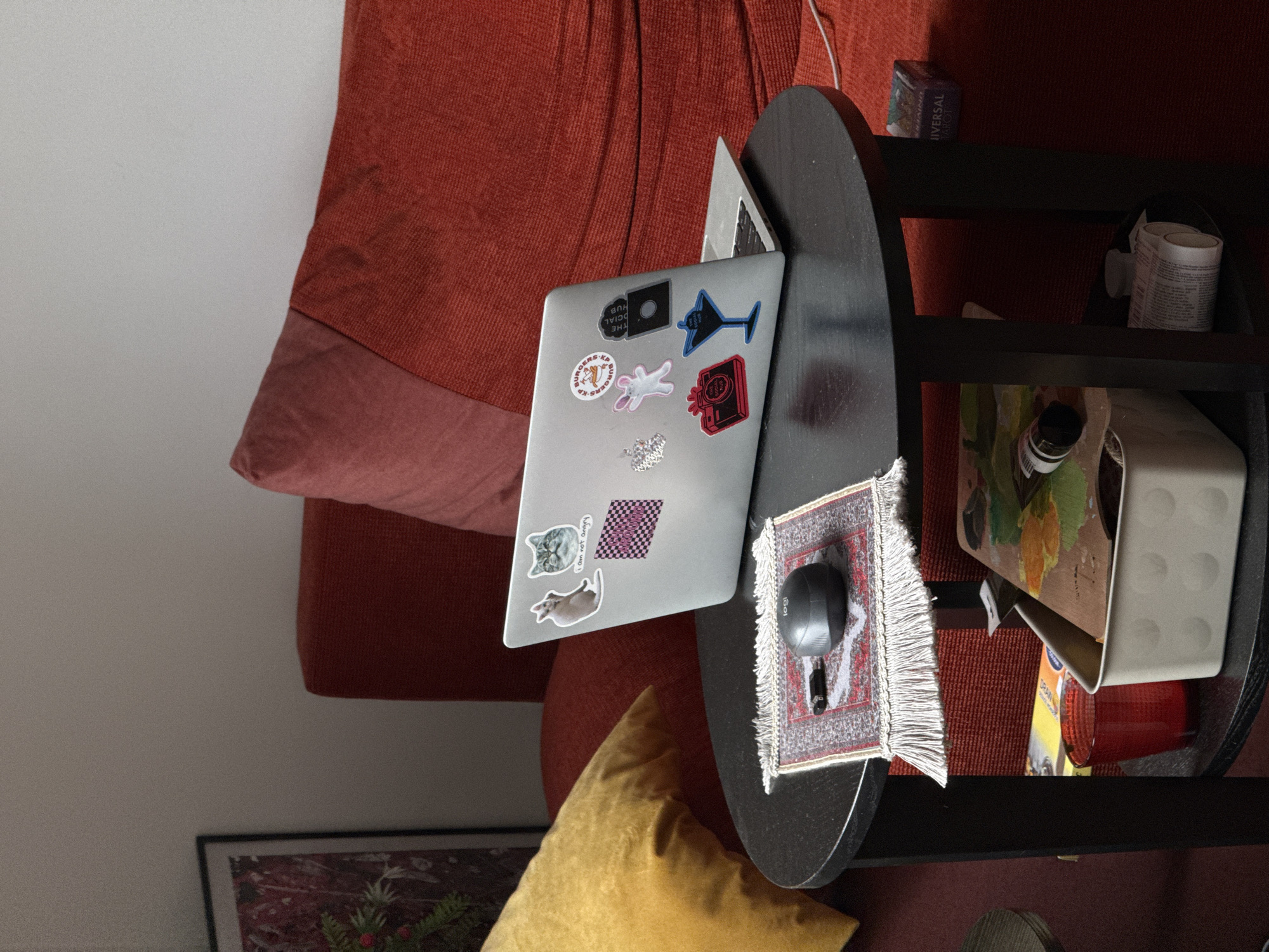 This image, taken in Vozdovac Urban Municipality, Serbia, presents an overhead view of an indoor setting, likely a living room or study corner, with a cozy and personalized atmosphere. A silver laptop, adorned with numerous stickers, rests closed on a dark, oval-shaped side table. Visible stickers include one with 'THE SOCIAL HUB', another with 'BURGERS K.P.' (partially visible and upside down), a blue martini glass, a pink bunny, a red camera icon (upside down), a grey cat head, a speech bubble reading 'I am not angry', and a pink and black checkered pattern.
Beside the laptop, a black Logitech computer mouse sits on a small, fringed rug with a traditional red and beige oriental pattern. The side table is part of a multi-tiered shelving unit, black in color. On the top shelf, a box labeled 'UNIVERSAL TAROT' is visible, along with a white cylindrical object containing unreadable text. A lower shelf holds a cream-colored container with dotted indentations, a red glass, a small bottle, and a piece of artwork featuring green and orange abstract elements.
The table is positioned next to a plush, red-orange corduroy-like sofa, which also features a lighter pink cushion or throw. On the floor below, a vibrant yellow throw pillow is visible, as is a portion of a framed picture displaying a reddish background with green foliage. No individuals are present, and the closed laptop suggests a moment of pause or an unoccupied space.