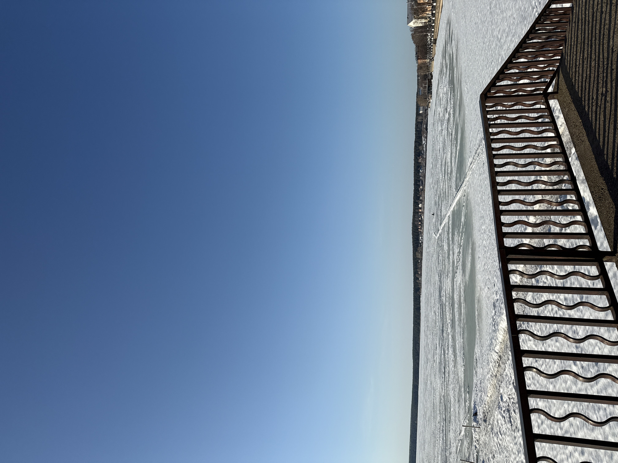 The image depicts a view from a bridge overlooking a frozen body of water, likely a river or lake, in Votkinsk, Russia. A metal railing with a wavy pattern is visible in the foreground on the right side of the frame. The ice-covered water extends towards the horizon, where a shoreline with trees and buildings is located. A clear, blue sky fills the majority of the frame on the left side. There are a few faint lines on the ice, possibly indicating skate tracks, and a single person is visible on the ice.
