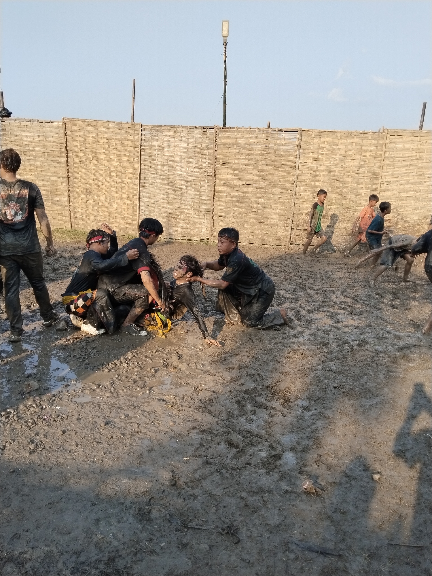 In a muddy area in Kedawung, Indonesia, several individuals are engaged in an apparent activity. A group of people, including adults and children, are covered in mud. Four adults are actively engaged in what looks like restraining another person, who is also covered in mud. Several children are running around in the background. The background consists of a woven bamboo fence and a light pole. The ground is entirely mud, suggesting a recent rain or a deliberately muddy area. The lighting suggests it is daytime.