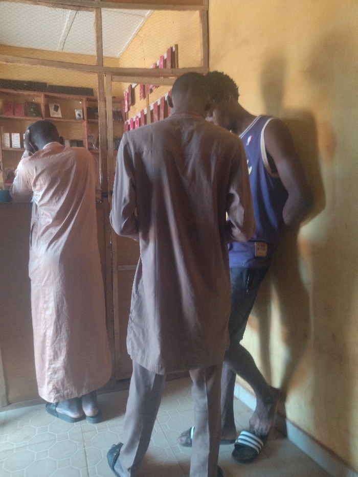 The image shows three men standing in a shop in Anyigba, Nigeria, located at coordinates 7.491, 7.175. The man on the left is wearing a long, brown robe and is leaning against a counter. The man in the center is wearing a brown shirt and pants and appears to be looking at something in the store. The man on the right is wearing a purple tank top and black shorts and is leaning against a wall. The shop appears to be a small, cluttered space with shelves on the back wall that are stocked with various items. The floor is tiled.