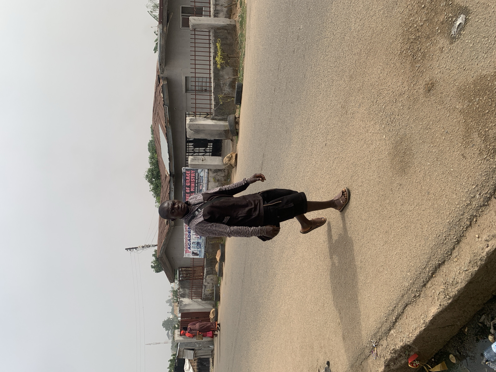 A medium shot depicts a man walking on a paved road in Jos, Nigeria. He is dressed in a dark jacket over a patterned shirt and dark shorts. The man wears leather sandals.
The road stretches into the background, flanked by buildings. A sign for “Precious Stone Academy Ministries” is visible on one of the buildings. Other people are present further down the road. The sky is overcast.