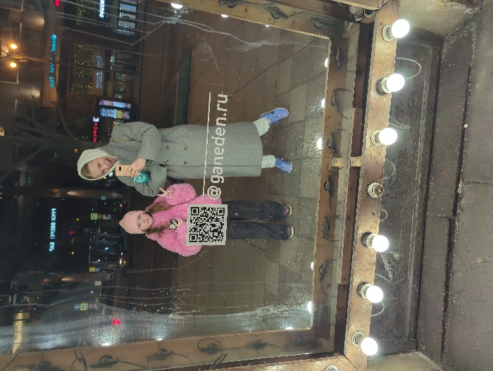 The image depicts a nighttime urban scene captured as a reflection in a vertical glass surface, which is framed at the bottom by raw wood planks featuring multiple exposed, glowing light bulbs.
**Scene Overview:** Two females, an adult and a child, are reflected, posing for a photograph. The background is a busy city street at night.
**People:**
* **Adult Female:** Appears Caucasian, in her late 20s to early 30s. She wears a long, light-grey double-breasted coat, light trousers, blue athletic shoes, and a light headscarf. She holds a light-colored smartphone with a teal case, angled as if taking the photograph, and has her mouth slightly open.
* **Child Female:** Appears Caucasian, approximately 7-10 years old. She wears a pink knitted beanie, a fluffy bright-pink jacket, dark patterned trousers, and dark shoes. She holds a dark smartphone in her left hand and extends her right hand, forming a "peace sign." She holds a white rectangular sign with a black QR code at chest level.
**Objects and Environment:**
* **Reflective Surface:** The glass surface exhibits smudges and visible water droplets. The text "@ganeden.ru" is clearly visible in white letters on the glass, above and to the right of the child.
* **Frame:** The lower edge of the reflective surface is bordered by a rustic, unfinished wooden frame containing at least six visible, illuminated spherical light bulbs.
* **Reflected Background:** The reflection reveals a dark urban environment at night. Prominent illuminated signs with reversed Cyrillic text are visible on distant buildings, including "РАБ ХРЕВВ КРАП" (reversed "ПАРК ВВЕРХ БАР"), "АПТЕКА" (Pharmacy), and "КОФЕ" (Coffee). Distant vehicle lights, streetlights, and multi-story buildings line the reflected street. The ground in the reflection appears to be paved with light-colored tiles.
* **Immediate Foreground:** Below the wooden frame, the ground consists of dark, rough concrete or asphalt.
**Location:** Based on the distinctive reversed Cyrillic signage "ПАРК ВВЕРХ БАР" (translating to "BAR UP PARK" or "PARK UP BAR"), the location is identified as Moscow, Russia, specifically near or at **Sadovnicheskaya Street, 62, building 5**, which is the address for "Бар Парк Вверх" (Bar Park Up).