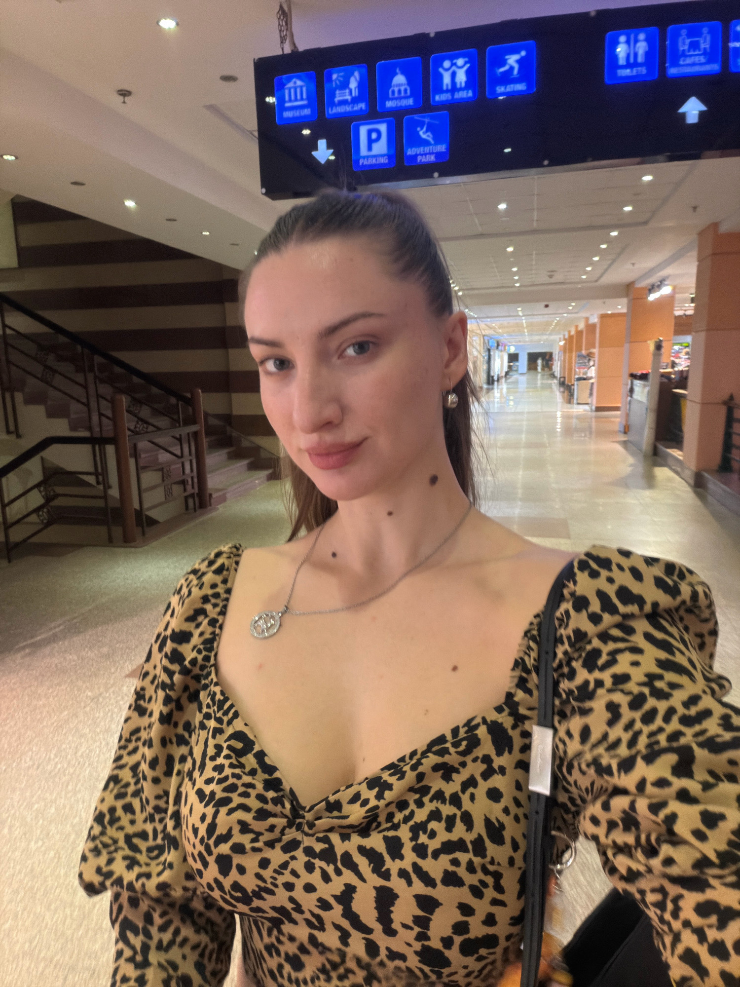 A young woman with brown hair pulled back in a ponytail takes a selfie in a well-lit indoor corridor. She is wearing a leopard-print top with puff sleeves and a silver necklace. A black strap of a bag is visible across her chest. In the background, there are escalators and a directional sign with icons for various facilities like "Museum," "Landscape," "Mosque," "Kids Area," "Skating," "Toilets," "Cafes," "Restaurants," "Parking," and "Adventure Park." The corridor appears to be in a commercial or public building, possibly an airport or a large mall, in Sharm El Sheikh, Egypt.