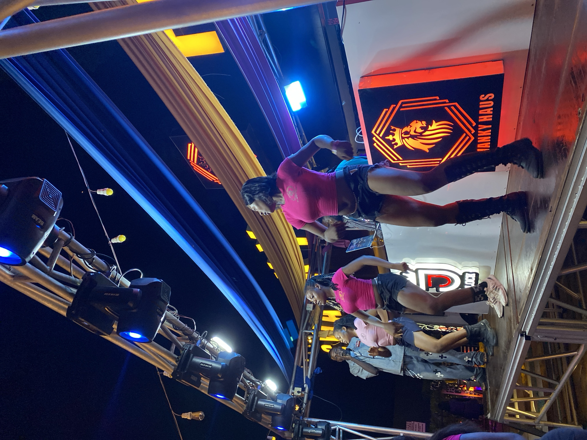 An outdoor night performance is taking place on a raised stage in Bar Jirgi Summa, Nigeria. Three young women are dancing on the stage, wearing vibrant pink tops and dark shorts. The woman on the right is wearing tall black boots, while the woman in the center is wearing athletic shoes. To the left of the dancers, a man is singing into a microphone, dressed in a striped shirt and jeans adorned with cross patterns. Behind the performers, a DJ setup is visible with a laptop. A large, illuminated sign featuring a lion's head and the text "Janky Haus" is positioned on the right side of the stage. The stage is lit by blue and yellow lights emanating from overhead fixtures, and the background is dark, indicating it is nighttime. The overall scene suggests a lively event or party.