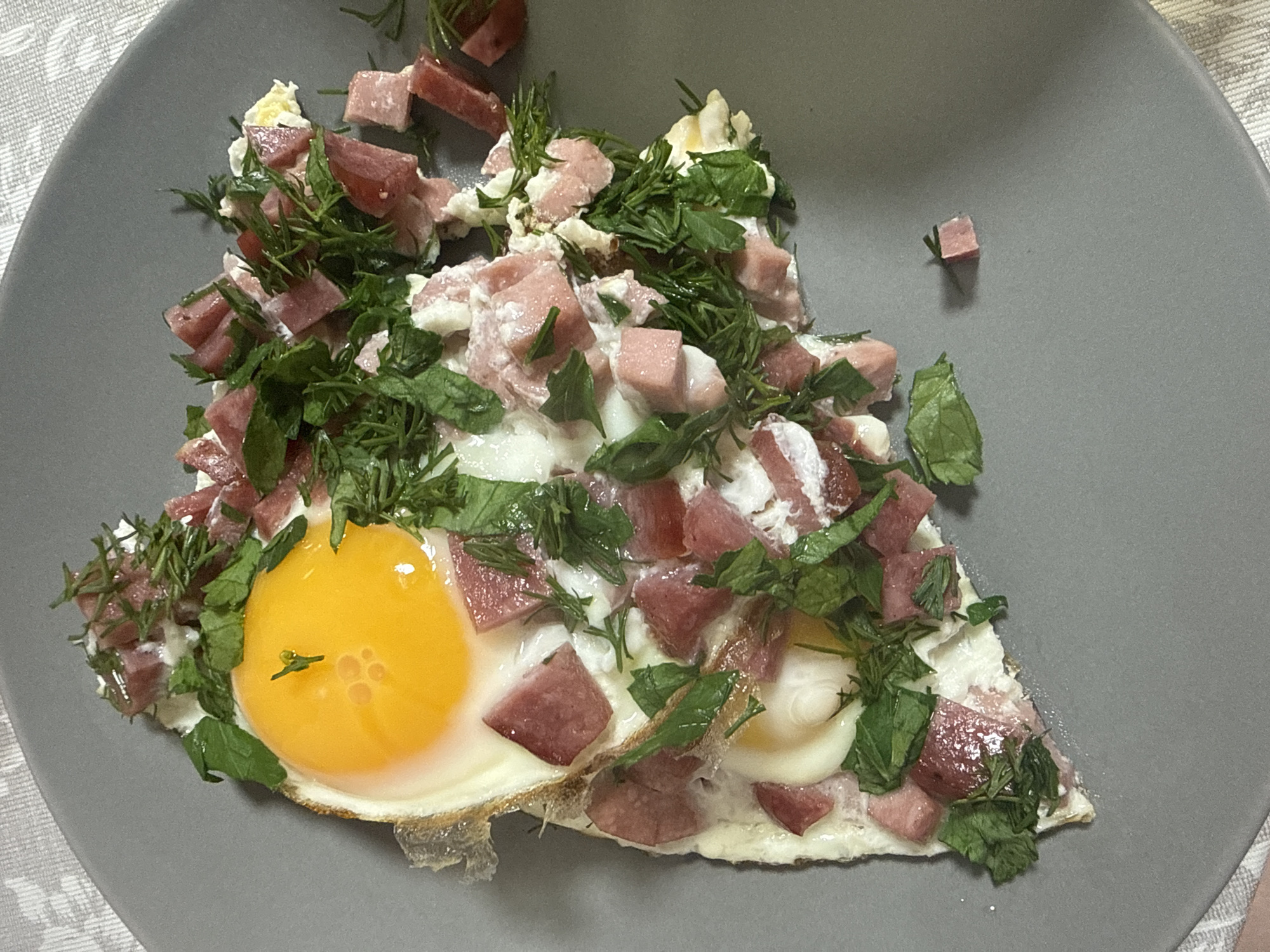 A close-up overhead shot presents a gray plate containing a slice of fried eggs topped with diced meat and herbs. The egg slice features two yolks, one of which is particularly prominent and bright yellow. The diced meat, cut into small cubes, is distributed across the egg white and between the herbs. The herbs appear to be a combination of dill and leafy greens. A single cube of meat sits separately on the plate. The background includes a hint of a textured surface, possibly a tablecloth or placemat. The image lacks locational context.
