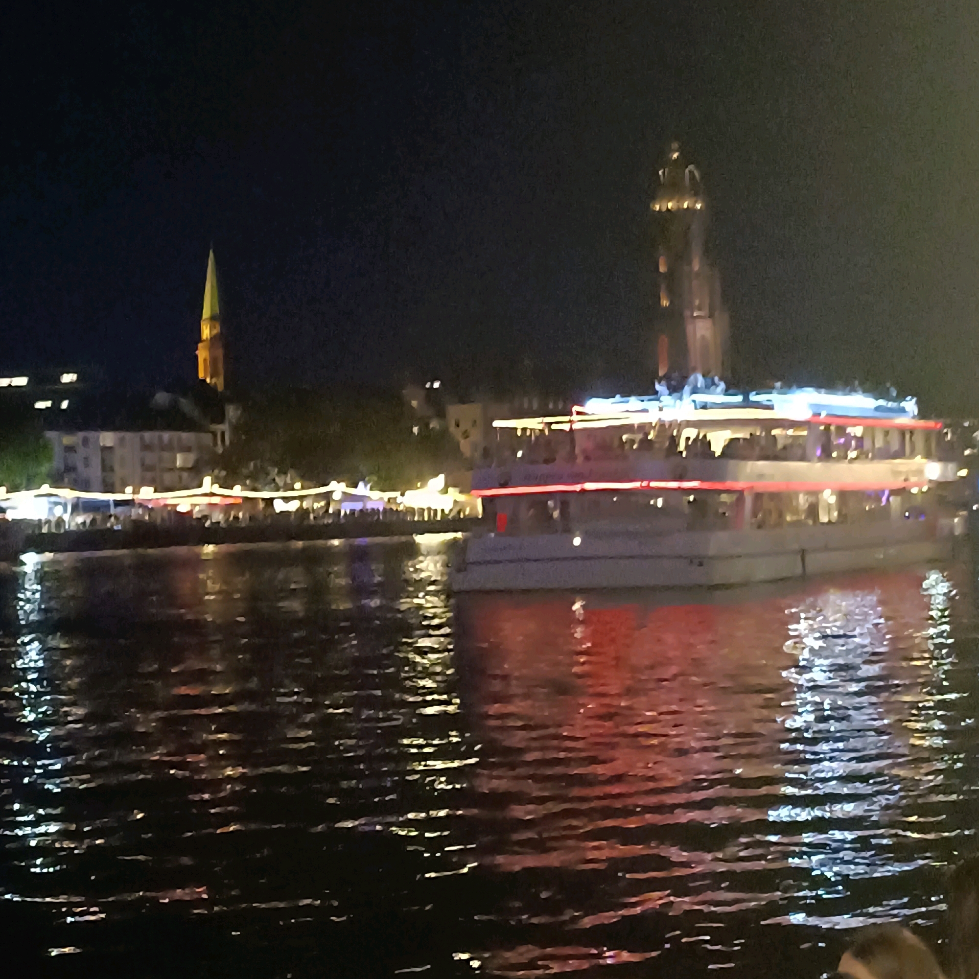 This is a nighttime photograph of the city of Frankfurt, Germany, likely taken from across the Main River. The scene depicts the illuminated skyline and a river cruise boat. In the background, a tall, spired church tower, possibly the Frankfurt Cathedral (Kaiserdom St. Bartholomäus), is visible, illuminated by yellow lights, with a green spire pointing towards the dark sky. To the left, other buildings are dimly lit.
In the foreground and middle ground, a large, multi-deck river cruise boat is prominently featured. The boat is brightly lit with red and blue lights along its sides and on its upper decks, where people can be seen gathered. The boat is navigating on dark water, reflecting the lights from the boat and the shoreline. Along the riverbank, there is a strip of illuminated tents or stalls, suggesting a lively promenade or market area with people. The water surface shows shimmering reflections of the lights, indicating movement and disturbance. The overall atmosphere is that of a vibrant city night scene along a riverfront.