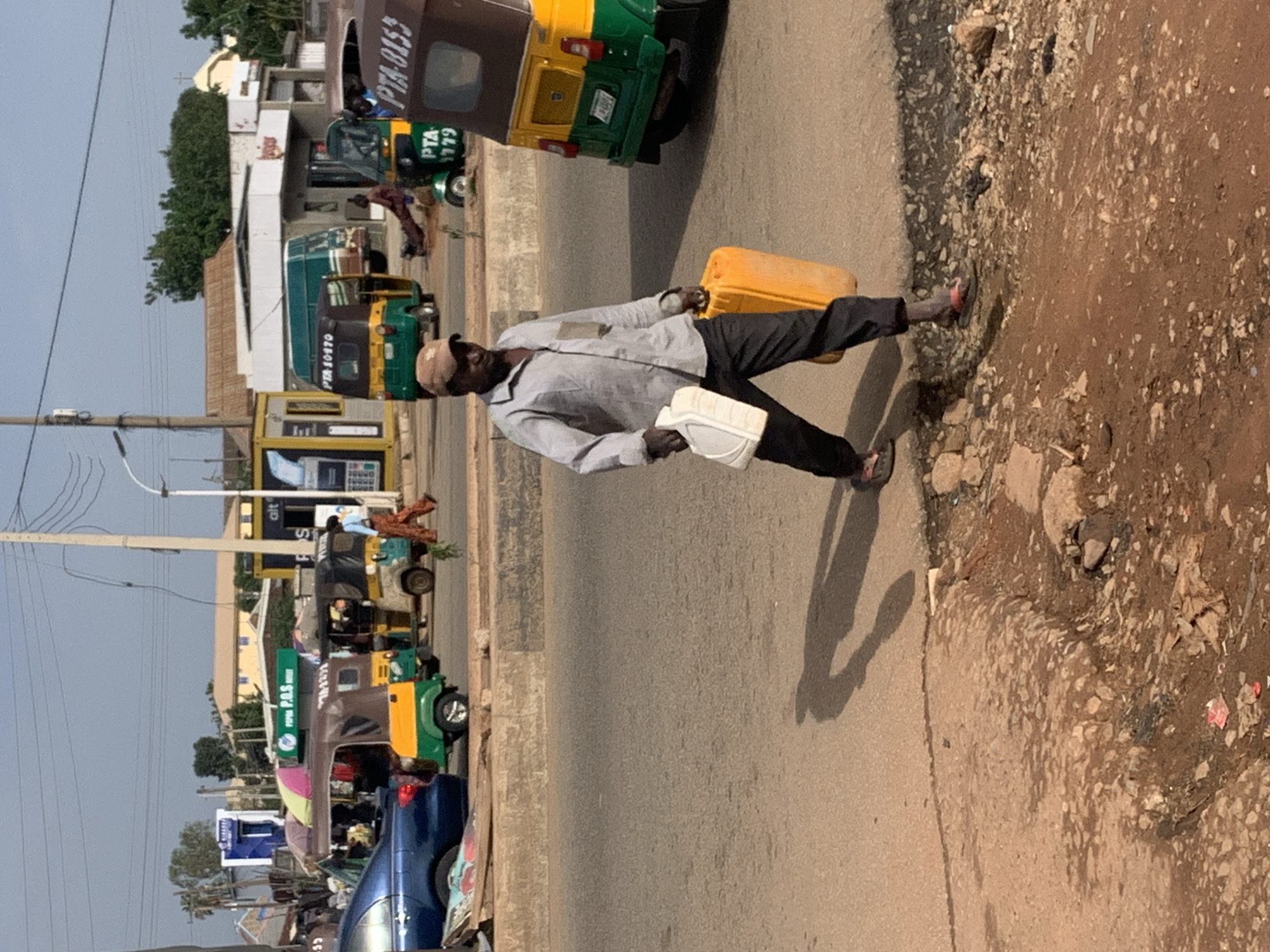 The image depicts a man walking on a dusty street in Gura Top, Nigeria, carrying two yellow jerrycans. He is wearing a light-colored, button-down shirt, dark pants, and sandals. The man is walking from right to left in the frame. Behind him, several vehicles are visible, including a blue car, a yellow and green auto-rickshaw, and a larger green and yellow vehicle. In the background, buildings and utility poles line the street. The ground is a mix of dirt and gravel, with some debris scattered around. The sky is visible in the upper portion of the image, appearing clear.