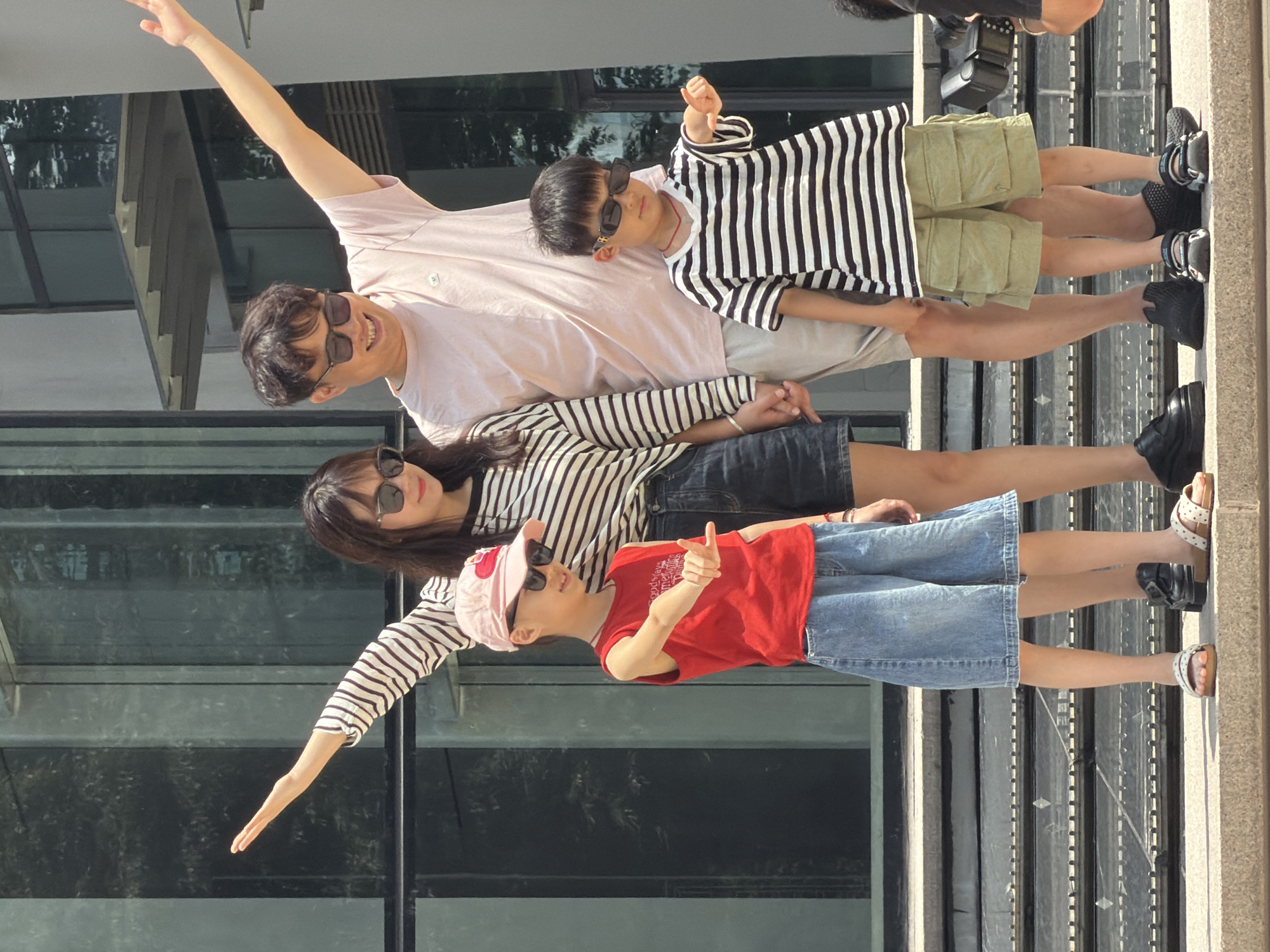 In Yuzhong District, China, a family of four, two adults and two children, poses for a photo on a sunny day. The family stands on a set of stone steps in front of a modern building with large glass windows. The adults, a man and a woman, are positioned behind the children. The woman wears a striped long-sleeved shirt and dark denim shorts. The man is in a light pink t-shirt and light-colored shorts. Both adults are wearing sunglasses.
The two children are in front. The child on the left is a boy wearing a red sleeveless shirt, denim shorts, a pink baseball cap, and sunglasses. He is extending his right arm. The child on the right is a younger boy wearing a striped t-shirt, khaki shorts with large pockets, and sunglasses. He is also extending his right arm. The family appears to be enjoying their time together.