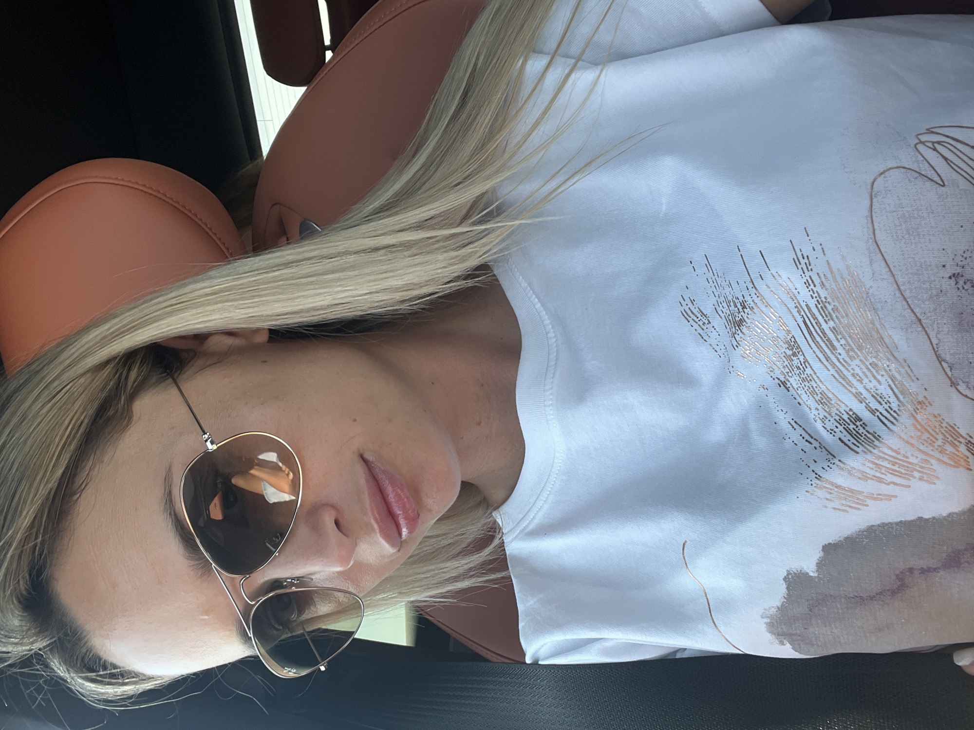 The image shows a close-up, waist-up shot of a blonde woman sitting in a car, likely in Sharm El Sheikh, Egypt. She is wearing aviator sunglasses with rose-gold frames and a white t-shirt with an abstract, metallic print. The car's interior is visible, featuring brown leather seats and a black headrest. The woman's hair is long and blonde, and she appears to be looking slightly down. Reflections of what appears to be interior car lights or the car's dashboard are visible in her sunglasses. The lighting suggests it is daytime.