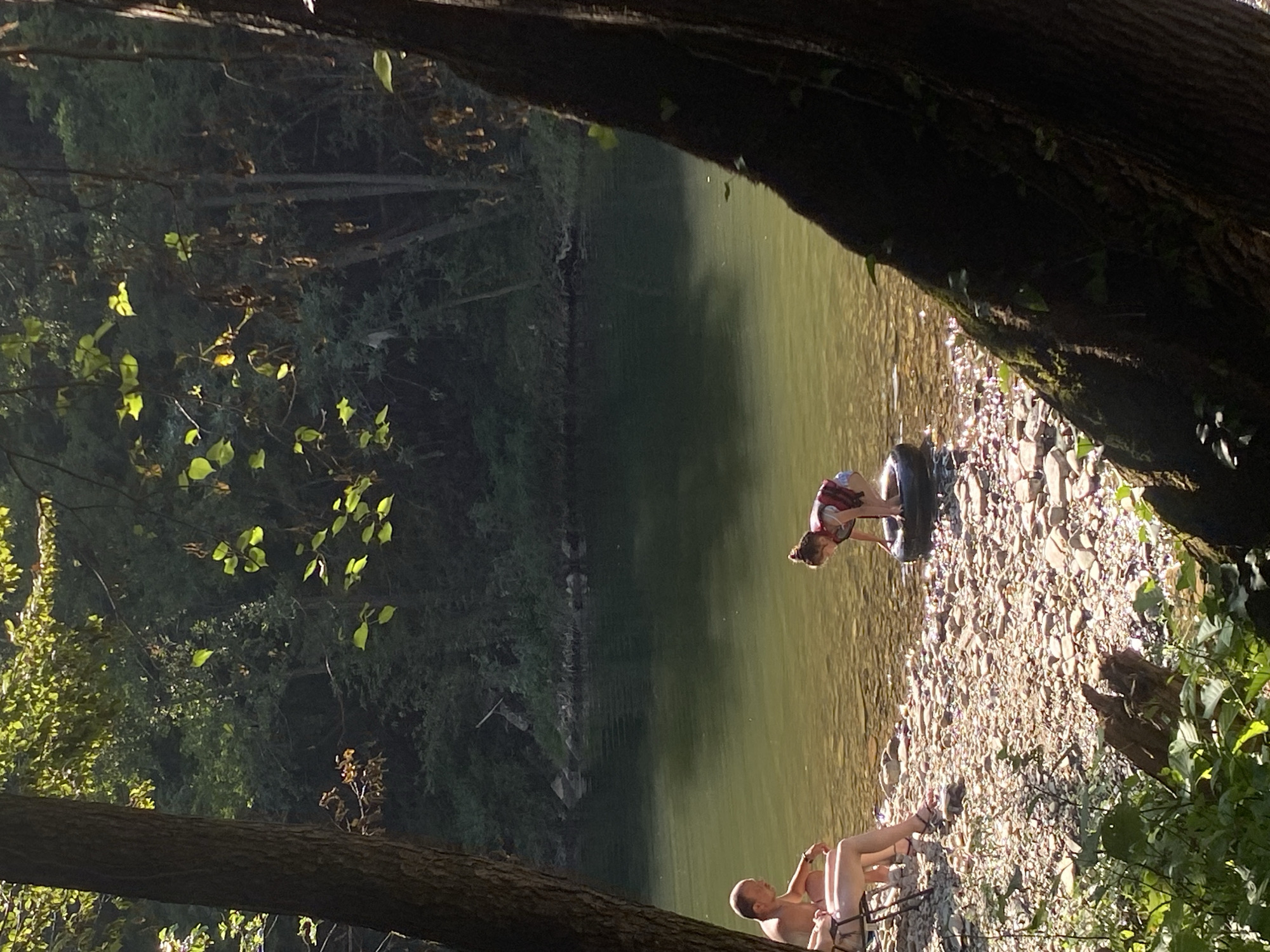 The image depicts a serene riverside scene, likely in or near Debnevo, Bulgaria, given the context. In the foreground, a person sits in a folding chair on a rocky shore, facing away from the camera towards the water. Further into the shallow water, a young child, wearing a life vest, is crouched inside an inner tube, appearing to play. The river is calm with a greenish hue, reflecting the surrounding lush, tree-covered banks. Sunlight filters through the foliage, creating dappled patterns on the water and shore. The atmosphere is peaceful and suggests a recreational outing by the river.