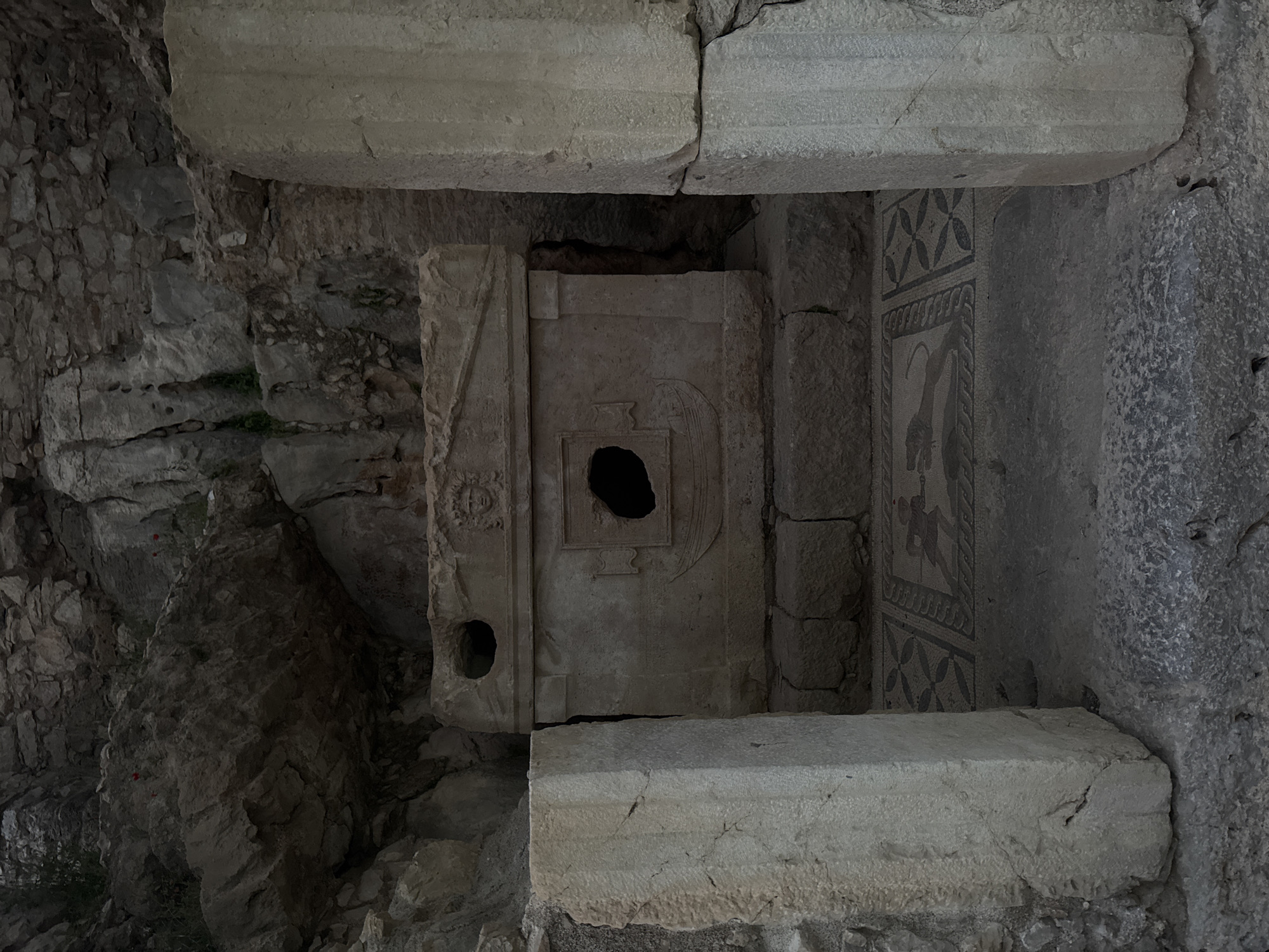 The image depicts an ancient stone tomb or rock-cut sanctuary, likely in Kumluca, Turkey. At the heart of the scene is a large, upright stone structure, resembling a sarcophagus, which is intricately carved. It features a prominent circular hole near its upper center and a smaller, similar opening towards its bottom-left side. Carvings on its surface include an inverted V-shape, possibly representing stylized wings or an eagle motif.
To the right of this central structure, a well-preserved mosaic panel adorns the wall. The mosaic exhibits geometric patterns, specifically repeating four-leaf clover or cross designs, at both its top and bottom sections. A central figural scene within the mosaic shows what appears to be a human or mythical creature interacting with an animal, rendered in dark tesserae against a lighter background.
The setting is an enclosed or semi-enclosed space, possibly a chamber or crypt, indicated by the large, cracked stone slabs forming the ceiling and a horizontal stone slab resting on the ground in the foreground. The left wall is constructed from rough, irregularly stacked stones with some small patches of green vegetation, while the right wall appears to be part of a natural rock formation, also containing a dark cavity. The overall lighting is dim, suggesting an internal or deeply recessed location. There is no discernible activity taking place, only the static display of ancient archaeological remains. No text is visible.