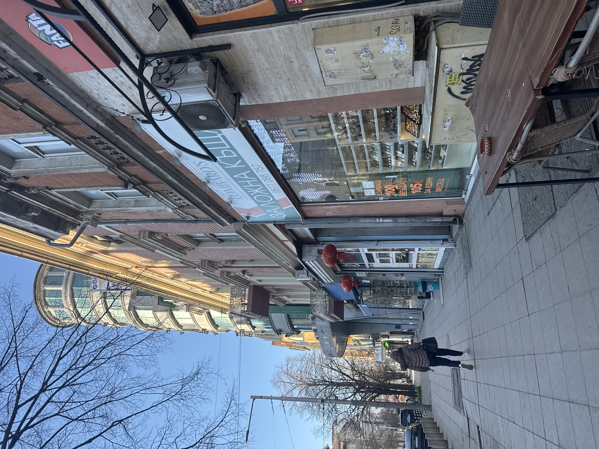 The image depicts a street scene in Sofia, Bulgaria. The view is angled upwards, showing buildings, trees, and the sky. A woman is walking on the sidewalk towards the lower part of the image. There are bare trees on either side of the street. Buildings line the street with a mixture of architectural styles and shopfronts. A shop sign visible reads "Lombard's," a pawnshop. There are cars parked along the street. The sky is blue and mostly clear.