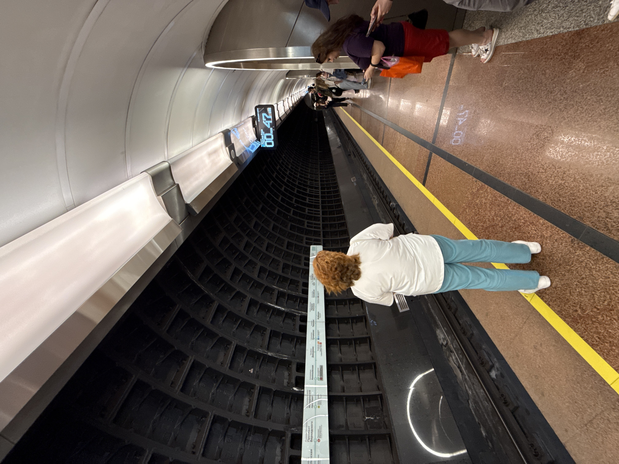 Here is a description of the image:
The image shows the interior of a metro station, possibly in Moscow, Russia, due to the Cyrillic script on signage. The camera is positioned at an angle. The walls of the tunnel where the tracks run are lined with dark, square panels. A woman in a white shirt and light blue pants is standing near the edge of the platform, looking at a vertical sign listing station names in Cyrillic. Further down the platform, there are a few other people standing. The floor of the platform is brown and marked with a yellow safety line. There is a digital clock on the wall, showing the time as "00:47." The overall environment appears modern and functional.