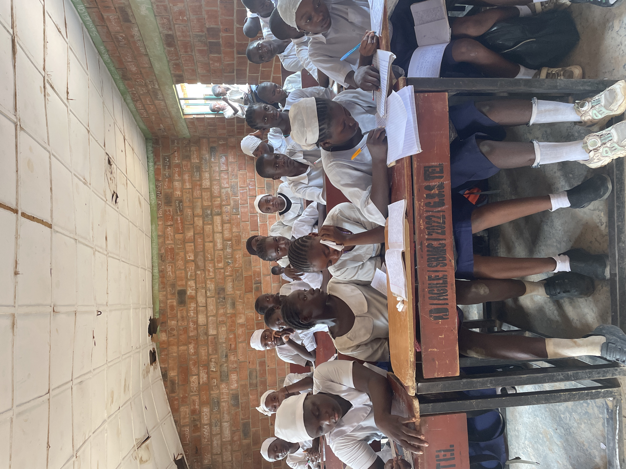 A classroom scene in Television, Nigeria, shows numerous young students, predominantly wearing white uniforms or head coverings, seated at wooden desks with metal frames. The students are engaged in an activity, with many holding pens and writing in notebooks, suggesting a lesson or exam is underway. Their gaze is generally directed forward, though some look towards the camera.
The classroom features rough brick walls, heavily adorned with handwritten names, messages, and graffiti in chalk or paint, including "Lord," "Mama Friga," "Favour ON GOD," "Bella Sal," "SLIVET David," and "SAMUEL HK CHAIRMAN SAMUELON." A section of the wall appears to be tiled or painted white with a grid pattern, also showing some wear and faint markings. Bright natural light streams into the room, likely from a window visible in the background, indicating daytime.
The wooden desks are sturdy and functional. One desk clearly displays the text "KD/AGILE/SBMC/2022/G.S.S. TEL.," with "G.S.S." commonly denoting Government Secondary School, and "TEL." aligning with the provided location context of Television. Students' legs and feet are visible beneath the desks, showing a mix of sneakers and socks, with some wearing dark blue shorts. The overall atmosphere suggests a focused learning environment in a school setting.