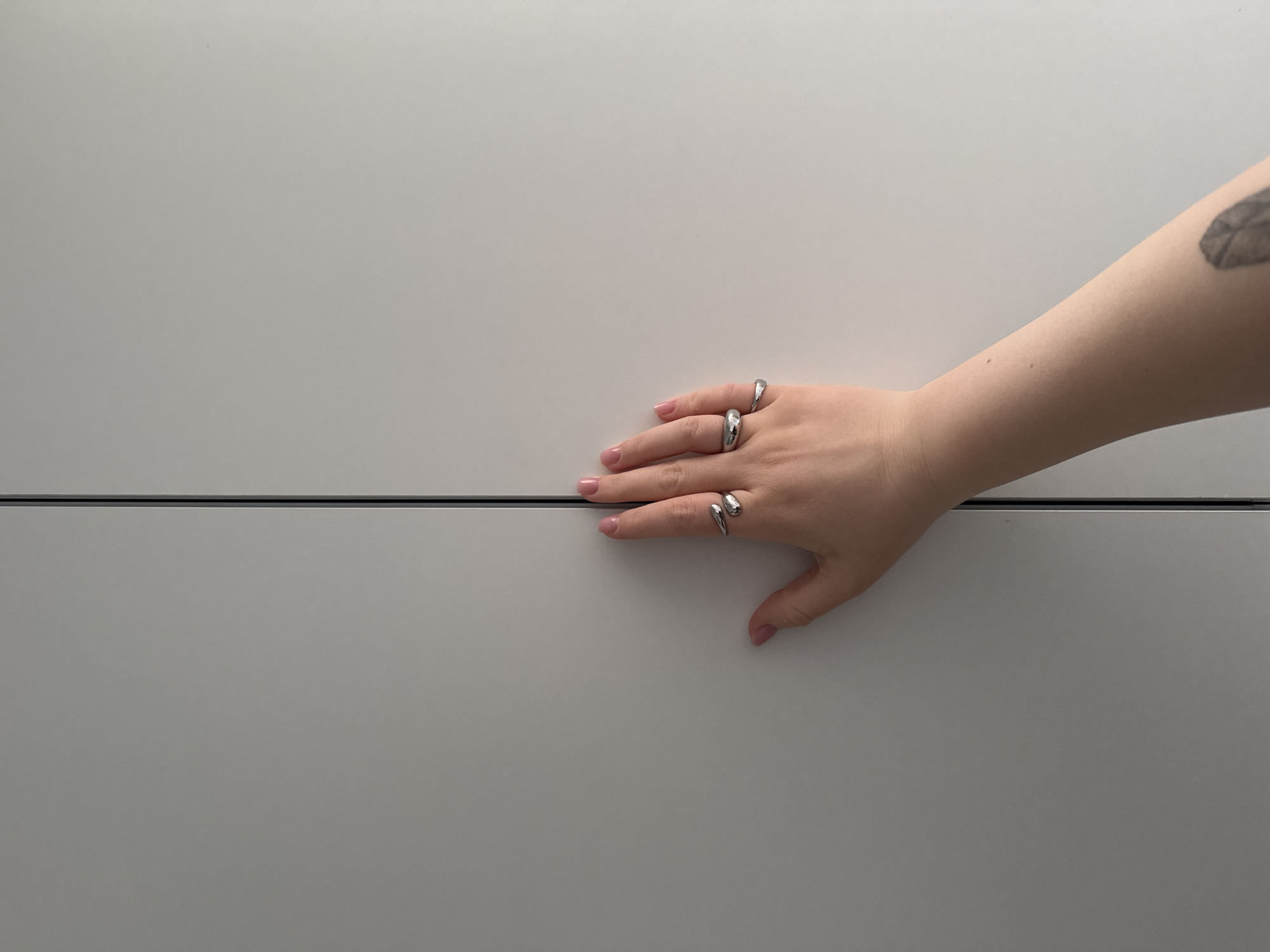 A person's arm and hand, adorned with three silver rings, rests on a light grey, two-panel wall. The arm, visible from the elbow down, has a tattoo near the elbow. The hand is positioned horizontally across the panels. The location is Unknown, Unknown.