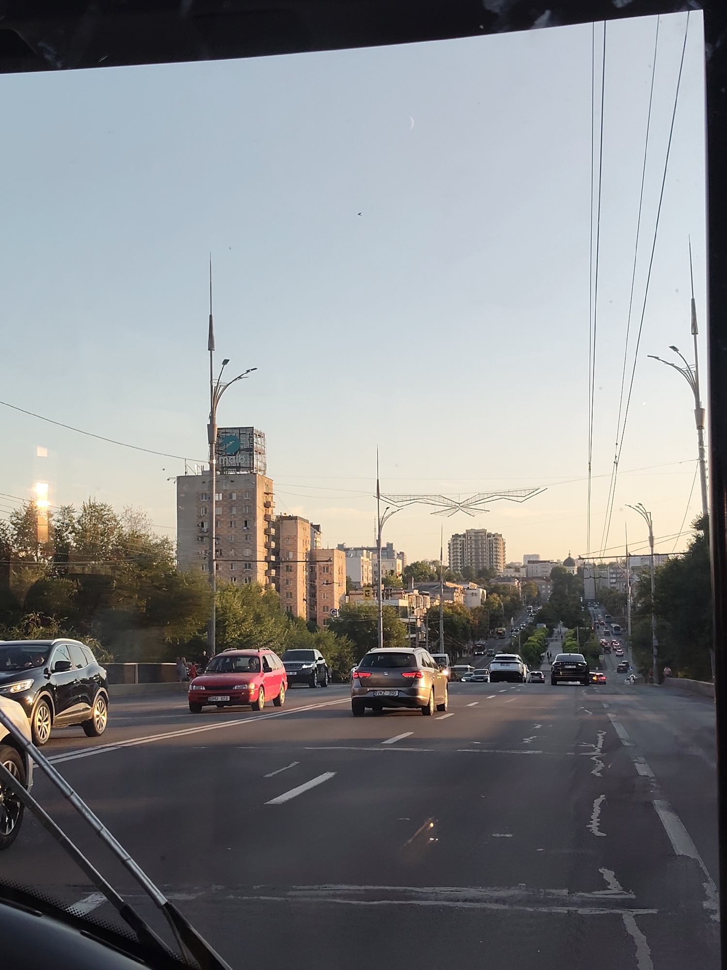 The image depicts a street scene in Chișinău, Moldova, likely during the late afternoon or early evening, given the golden hue of the sunlight. In the foreground, a busy multi-lane road is visible, with several cars in motion. A black SUV is in the left lane, a red hatchback is in the middle lane, and a silver sedan is further down the road. The road is marked with white lines indicating lanes and crosswalks.
The middle ground features a cityscape with a mix of residential and commercial buildings. Tall, multi-story buildings, some with balconies and many windows, dominate the skyline. A prominent feature is a tall apartment building with a billboard-like structure at its top, displaying a green logo. Trees and other vegetation line the sides of the road and are interspersed between the buildings, suggesting a degree of greenery within the urban environment. Streetlights and overhead power lines are visible, indicating the city's infrastructure.
In the background, the road continues uphill, lined with trees and more buildings. A distant view shows a long line of cars traveling up the incline, suggesting a significant traffic flow. The sky is a clear, pale blue, with a faint crescent moon visible in the upper left quadrant. The overall impression is of an active urban environment with the characteristic architecture and infrastructure of a Eastern European city. The perspective suggests the photo was taken from within a vehicle, possibly a bus or car, looking forward.