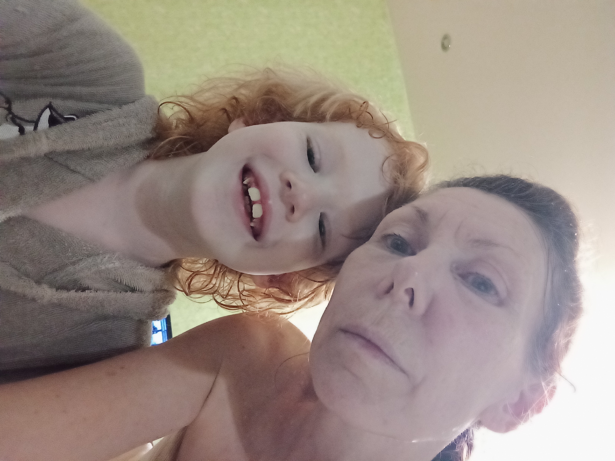 This is a selfie taken from a low angle, looking upwards at two subjects. The subject in the foreground and on the right is an adult woman, with fair skin and dark hair pulled back. Her expression is neutral, with her eyes looking forward. The subject in the background and on the left is a young child with red, curly hair and fair skin. The child is smiling broadly, showing teeth. Both subjects appear to be wearing bathrobes. The setting appears to be indoors, possibly a bathroom, with a green-tinted wall visible behind the child and a lighter, beige wall or ceiling behind the adult. The lighting is bright and appears to be coming from above and to the right, creating some glare. The overall impression is of a casual, intimate moment captured between the two individuals. There is no visible text in the image.