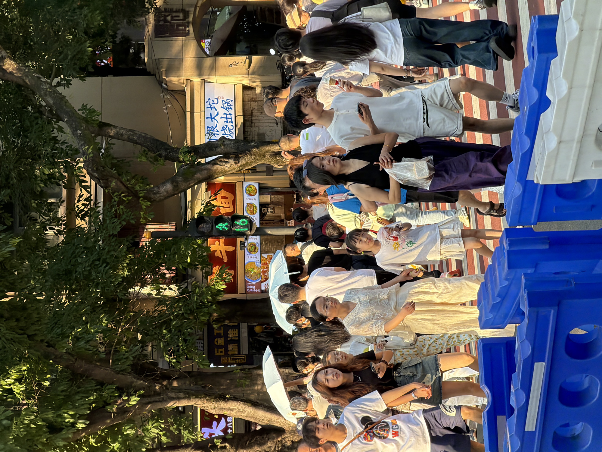 In Yuzhong District, China, a pedestrian crossing is occupied by a crowd of people. The sun is casting long shadows, indicating it is likely late afternoon or early morning. Pedestrians are dressed in casual summer attire, including t-shirts, shorts, and skirts. Some are holding umbrellas for shade. Traffic signals are visible in the background, and signage in Chinese characters suggests commercial establishments. Blue and white barriers are present on the street, potentially indicating a temporary closure or construction. The scene depicts a typical urban environment with daily life unfolding.