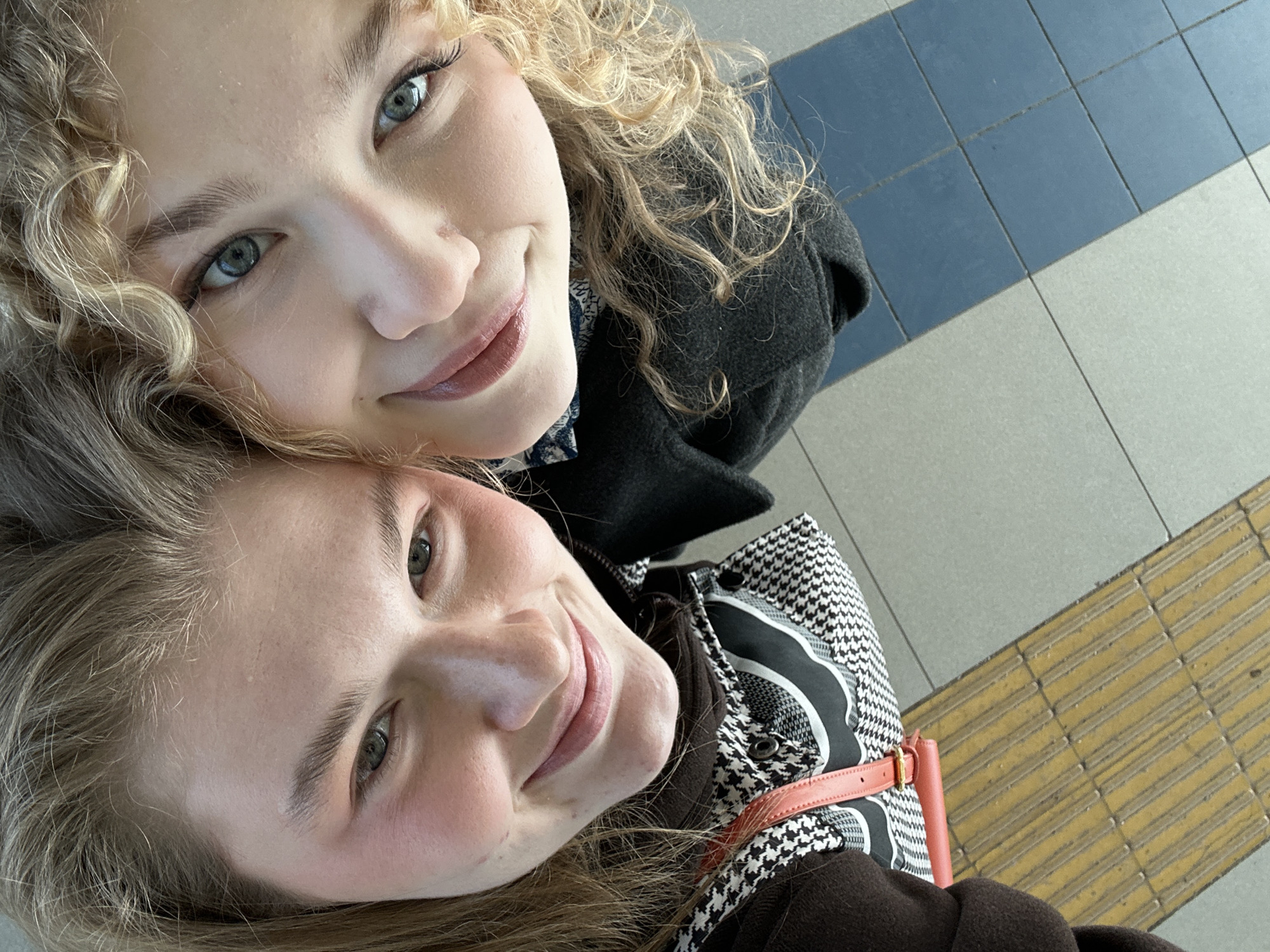Two young adult females are depicted in a close-up, upward-angled view, consistent with a selfie, within an indoor or covered outdoor public space in Vorob'evka, Russia. The female on the left has light brown hair, green eyes, and light skin, displaying a subtle smile. She wears a dark brown outer garment over a black and white houndstooth patterned layer, with a coral-colored strap visible over her shoulder. The female on the right has curly blonde hair, blue eyes, and light skin, exhibiting a smile with slightly parted, glossy lips. She is dressed in a dark grey or black coat over a blue and white patterned garment. Their faces are in close proximity, indicating a shared, friendly interaction. The floor visible in the lower portion of the image consists of a strip of yellow tactile paving adjacent to light grey tiles, which transition diagonally into an area of dark blue tiles.