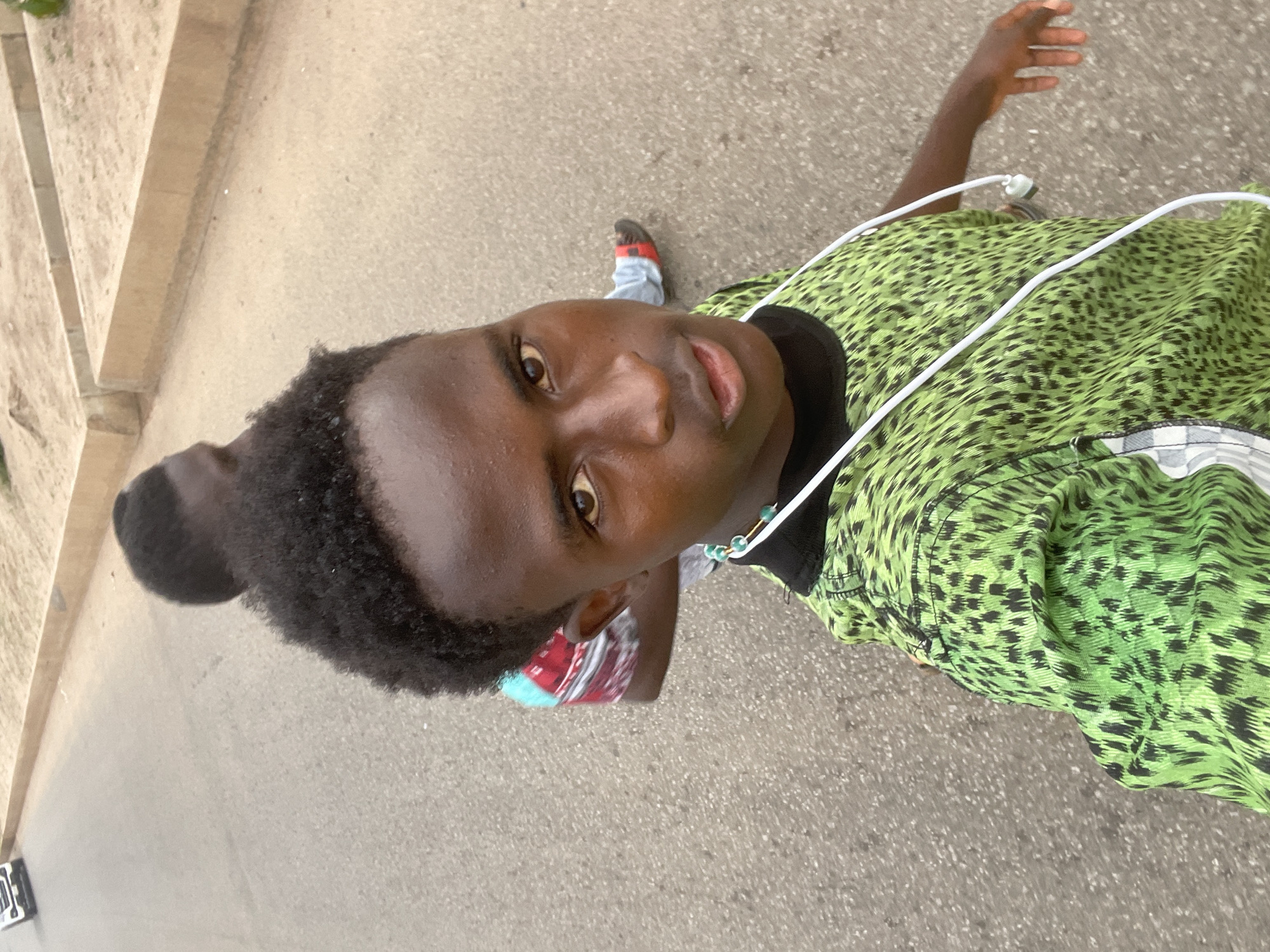 The image shows two young men walking on a paved road, likely in Jos, Nigeria. The person in the foreground is taking a selfie, facing the camera. They have dark skin, short curly hair, and are wearing a vibrant green t-shirt with a black and green leopard-print pattern. A white cord for earbuds is draped around their neck and over their shirt. They are also wearing a beaded necklace with white, blue, and gold beads.
The second man is walking behind the first, his back mostly to the camera. He is wearing a shirt with a red, blue, and white pattern and what appear to be jeans and sandals. The road is asphalt, with a concrete curb and sandy embankment visible on the right side. In the background, a white SUV is driving away on the left side of the road. The overall lighting suggests it is daytime, possibly late afternoon.