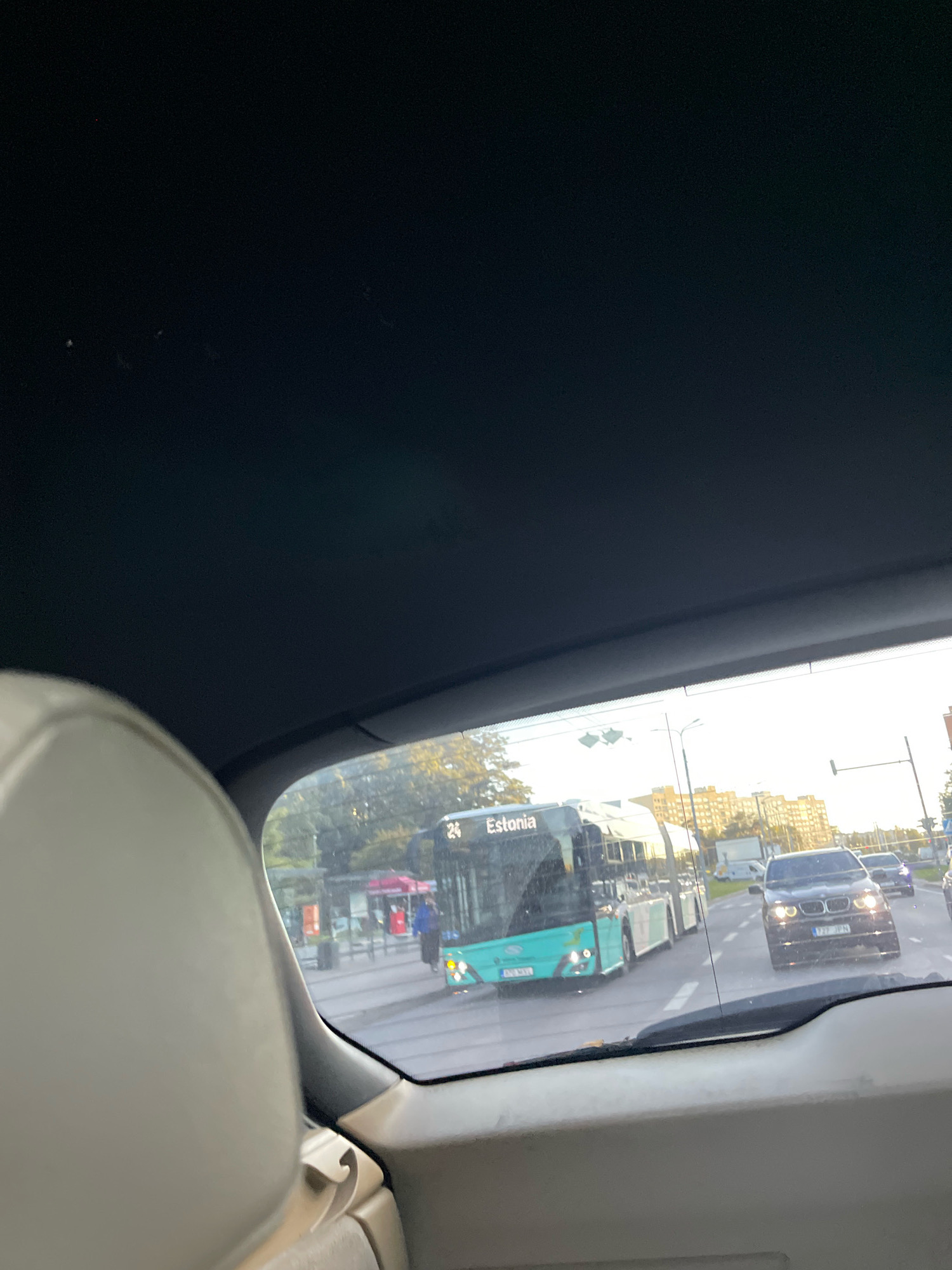 This is a view from inside a vehicle, looking out through the rear windshield. The perspective shows the road ahead and the surrounding environment in Tallinn, Estonia. A light-blue and white bus, with "Estonia" written on its side and the number "24" displayed, is prominently in the left lane. To the right of the bus, a black BMW sedan with its headlights on is in the right lane. Further ahead, other vehicles and buildings are visible, including a row of apartment buildings with yellow facades. The sky is bright, suggesting it is daytime, likely late afternoon given the warm sunlight casting long shadows. Traffic lights are visible above the intersection ahead. There are indications of a bus stop area on the left side of the road with people and covered shelters. The interior of the vehicle is partially visible in the foreground, including a light-colored seat.