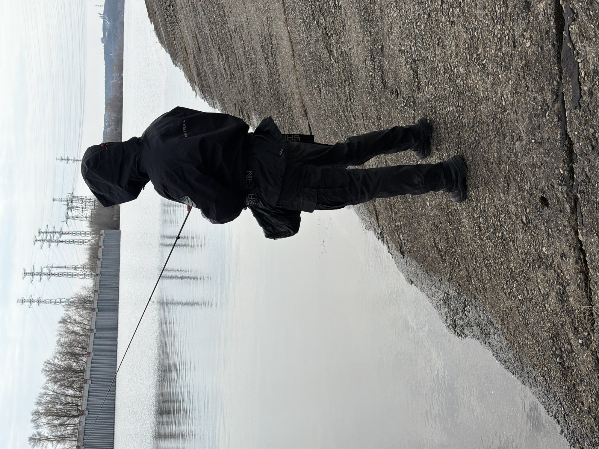 A person wearing a black jacket, pants, and shoes stands on a paved surface next to a body of water, holding a fishing rod. The water reflects the sky and nearby objects. Power lines and trees are visible in the background. The location is Chaikovsky, Russia.
