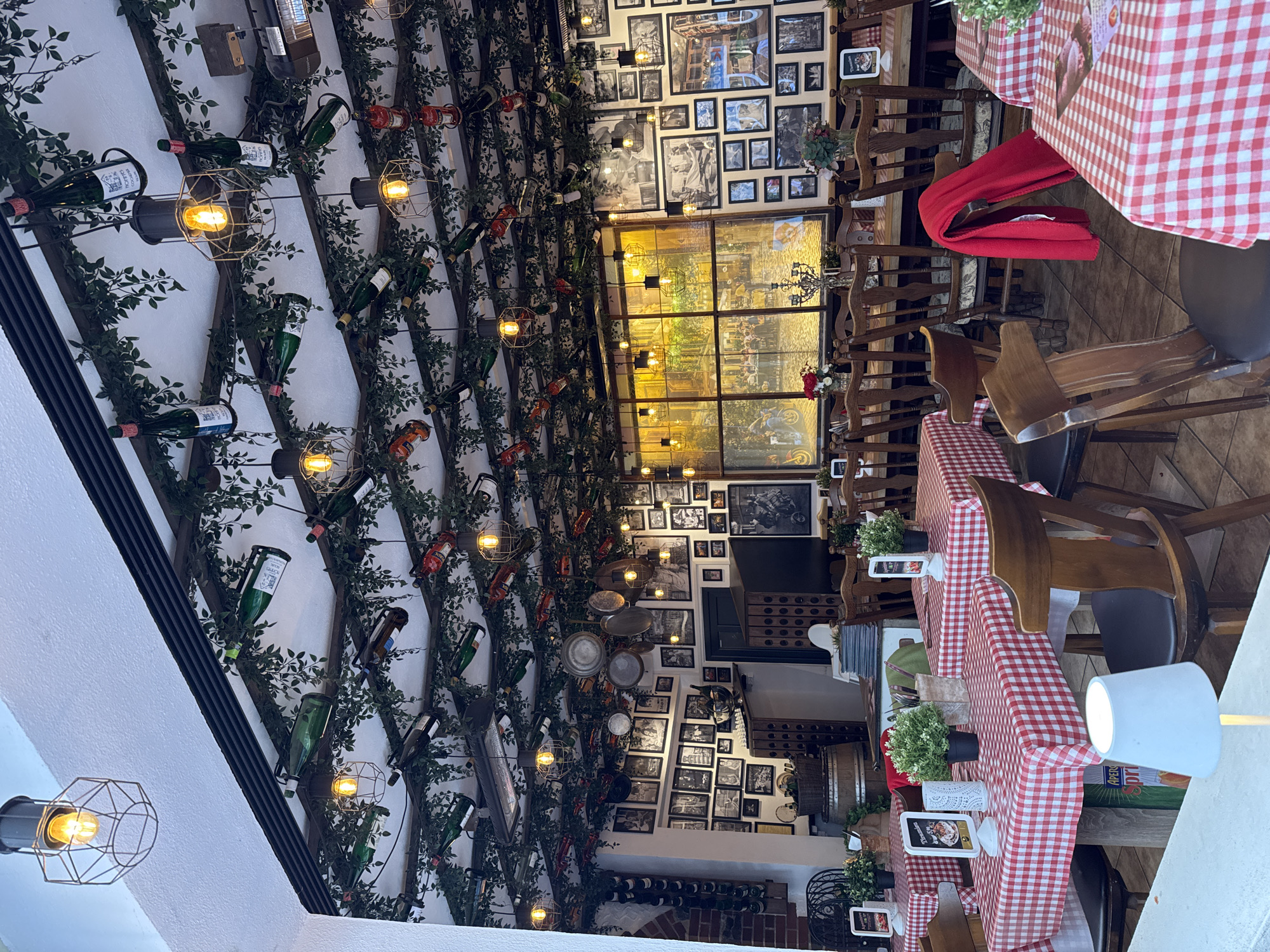 The image depicts the interior of a restaurant in Cochem, Germany. The decor includes wine bottles and faux greenery arranged on the ceiling, creating a vineyard-like ambiance. The walls are adorned with numerous framed photographs. Tables are set with red and white checkered tablecloths. The restaurant features wooden chairs and lighting fixtures with exposed bulbs. There are no people visible in the frame.