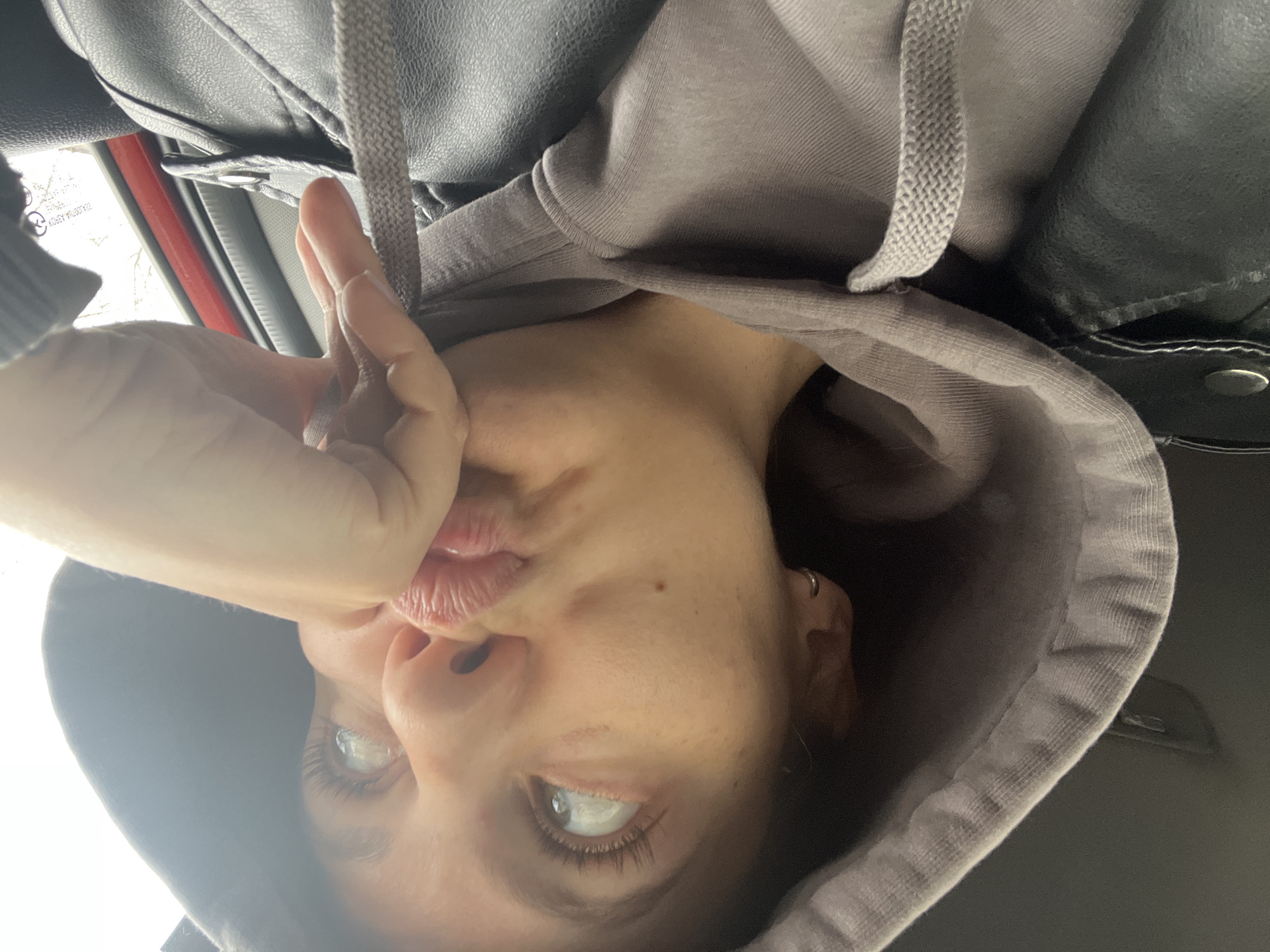 The media file, captured in Chaikovsky, Russia, features a close-up selfie of a person, rotated 90 degrees counter-clockwise from a standard upright orientation. The individual, seen from the chest up, is wearing a grey hooded sweatshirt underneath a dark, possibly leather, jacket.
The person is engaged in a playful activity, holding the grey drawstring of their hoodie with their fingers near their mouth, which is puckered into a kissing or cheeky expression. Their eyes are open, looking directly at the camera, with visible long eyelashes and bright reflections suggesting a strong light source. A small, simple hoop earring is visible in their left ear (on the right side of the rotated image). A small mole or blemish is present on their right cheek.
The background suggests the interior of a vehicle, likely a car, with a bright window area indicating daylight outside. A distinct red trim runs along what appears to be the car's window frame. Faint, mirrored text "KOTSEY PLO039Z," possibly a reflection of a license plate or vehicle identification number, is visible on the window surface. The overall mood of the image is casual, playful, and informal, suggesting a daytime setting.