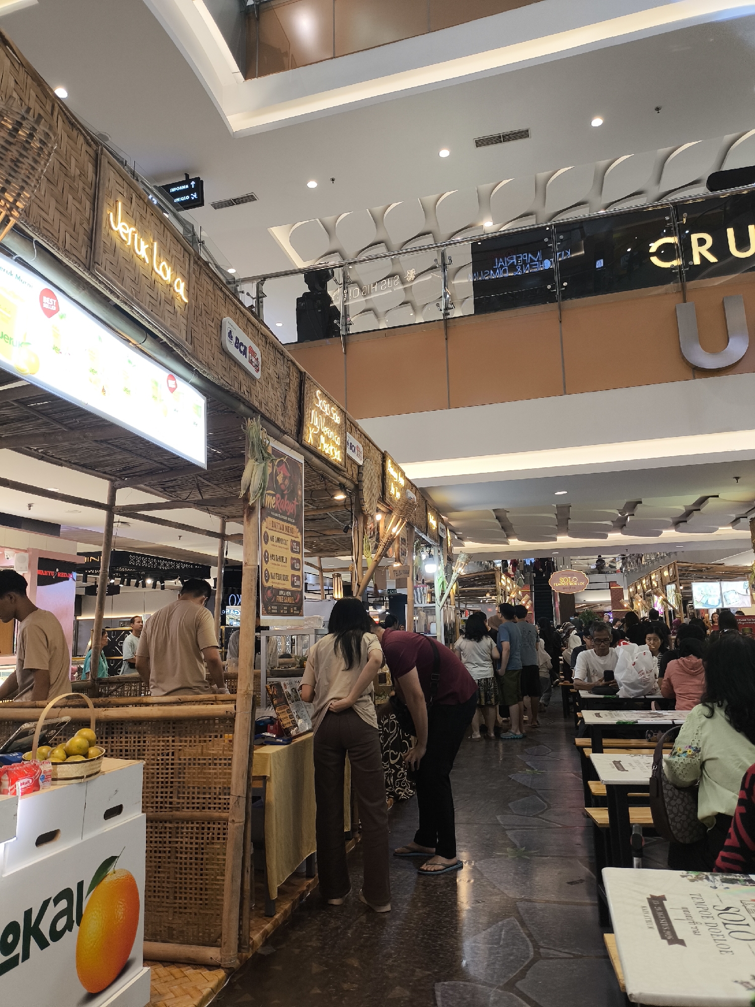 The image depicts a bustling food court or market area, likely in the city of Grogol, Indonesia. The architecture suggests an indoor shopping mall with high ceilings and decorative paneling. In the foreground, a food stall is visible, constructed with bamboo and straw, featuring a sign with neon lettering that reads "Jeruk Lava". Next to it, there are boxes with the branding "lokal" and an illustration of an orange. Several people are present, including vendors and customers. One vendor, wearing a light brown shirt, is standing behind the stall. Customers are seen browsing, ordering, and eating at tables. Further in the background, more food stalls and dining areas are visible, with a diverse crowd of people interacting. Signs for various businesses, including "CRU U", are present on the upper levels. The overall atmosphere is lively and commercial, indicative of a popular public gathering space.