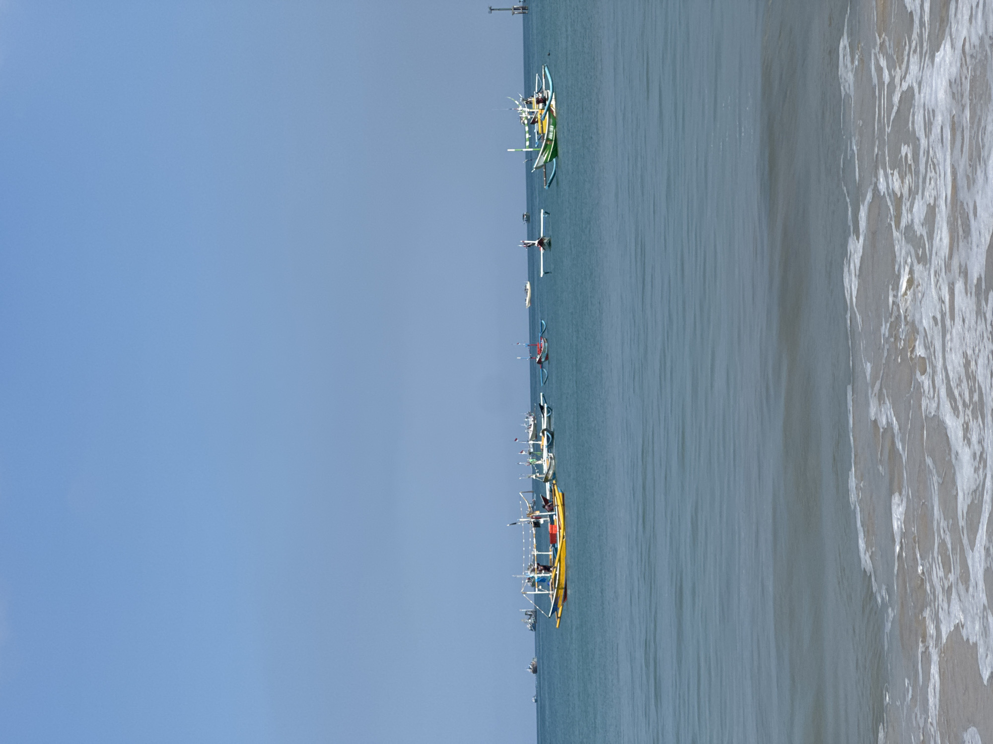 This media file captures a serene coastal scene in Kuta, Indonesia, under a clear, bright blue sky. The image primarily features a wide expanse of ocean, with several traditional Indonesian outrigger fishing boats, known as Jukungs, lined up across the calm waters.
The sky, occupying the upper portion of the image, is a uniform, light blue, suggesting clear weather conditions. Below the sky, the ocean stretches out, displaying varying shades of blue and green, indicating a relatively calm sea surface further from the shore.
A notable array of Jukungs are visible, distinguished by their colorful hulls and characteristic outriggers designed for stability. One prominent yellow Jukung is positioned closer to the viewer, while other boats, including a green and white one, are dispersed further along the watery expanse. The boats appear to be either anchored or moving slowly, possibly engaged in fishing activities or simply resting near the coast. No specific individuals are clearly discernible on the boats.
Towards the right side of the image, the scene transitions to the sandy beach, where gentle white foamy waves are breaking. The texture of the water in this area shows the movement and froth of the waves washing ashore.
The overall impression is one of peaceful daytime, likely morning or early afternoon, with calm weather. There is no visible text in the image.