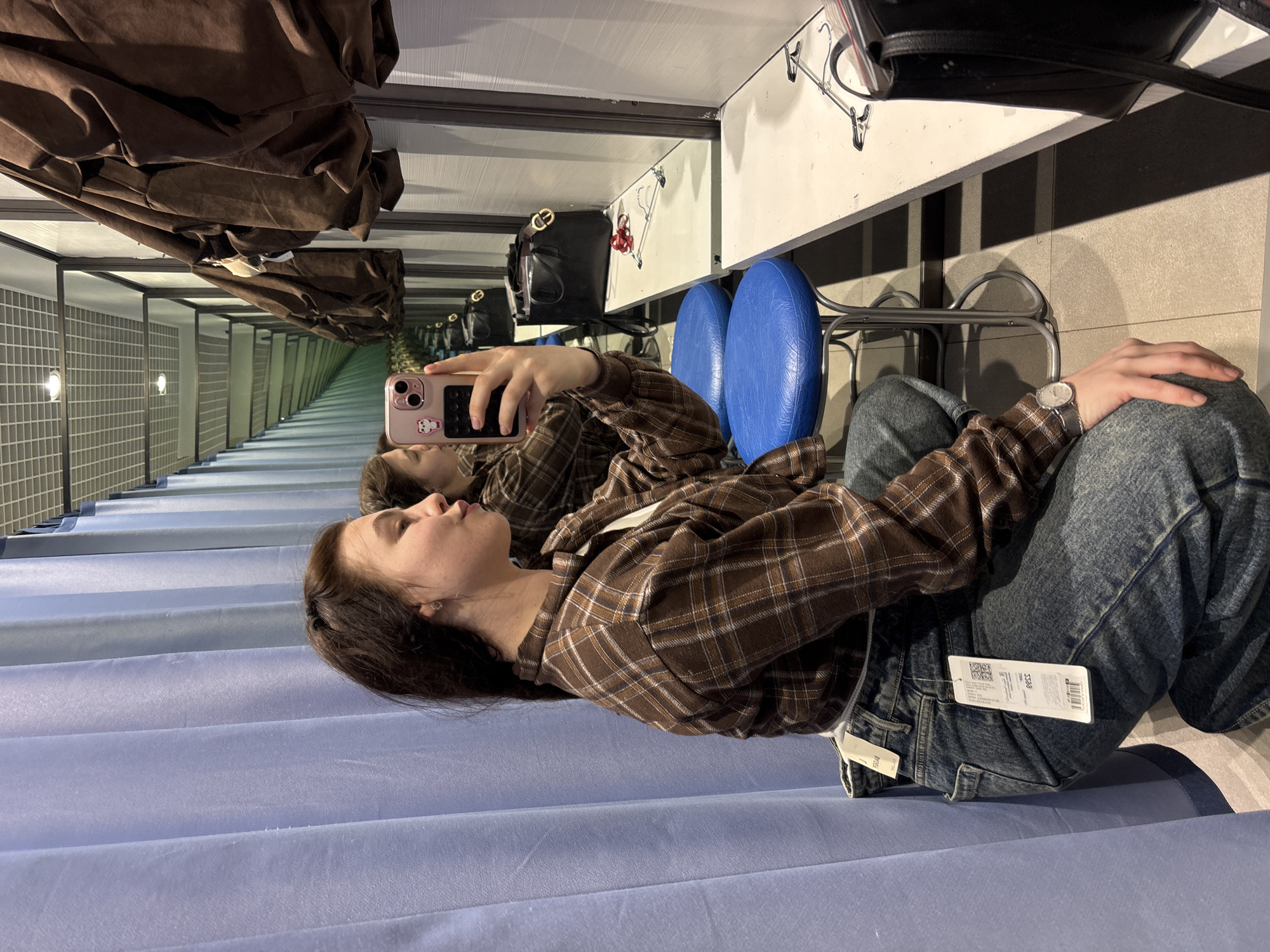 A young person is depicted taking a mirror selfie while seated in what appears to be a changing room of a retail store. The person, likely female, is wearing a brown and white plaid long-sleeved shirt over a white undershirt, paired with blue denim jeans that still have price tags attached. A silver-cased watch with a dark strap adorns their left wrist. They are holding a light pink smartphone with a distinctive white character case (resembling a bear or similar animal) in their right hand, angled to capture their reflection.
The setting is characterized by light blue fabric changing room curtains to the person's left, and a white shelf or counter to their right, upon which a black handbag rests. Two blue, oval-shaped stools are visible near the person. The mirror prominently reflects a long, receding corridor lined with identical changing room partitions and grid-patterned walls, creating an illusion of depth and suggesting a spacious store environment. Dark brown clothing items are draped overhead, also reflected.
The activity taking place is a self-portrait taken within a retail changing area, likely during a shopping trip. The person appears calm and focused on the task. Notable visible text includes "eess", a QR code, and "SM 28" printed on the white tags attached to the jeans. The time of day is not indicated, but the scene is indoors under artificial lighting.